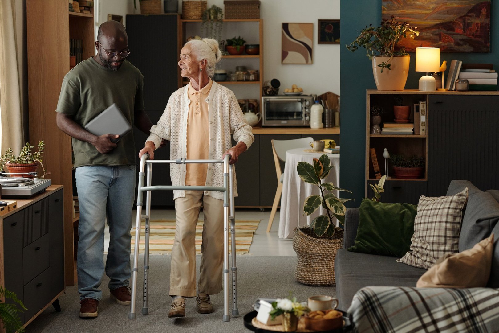 Senior Caucasian woman using walker for support walking indoors accompanied by Black man holding laptop, both interacting and smiling in modern home living space