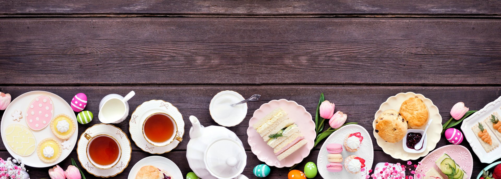 Easter or spring tea bottom border against a dark wood banner background. Assortment of finger sandwiches, scones and sweet desserts. Top down view. Copy space.
