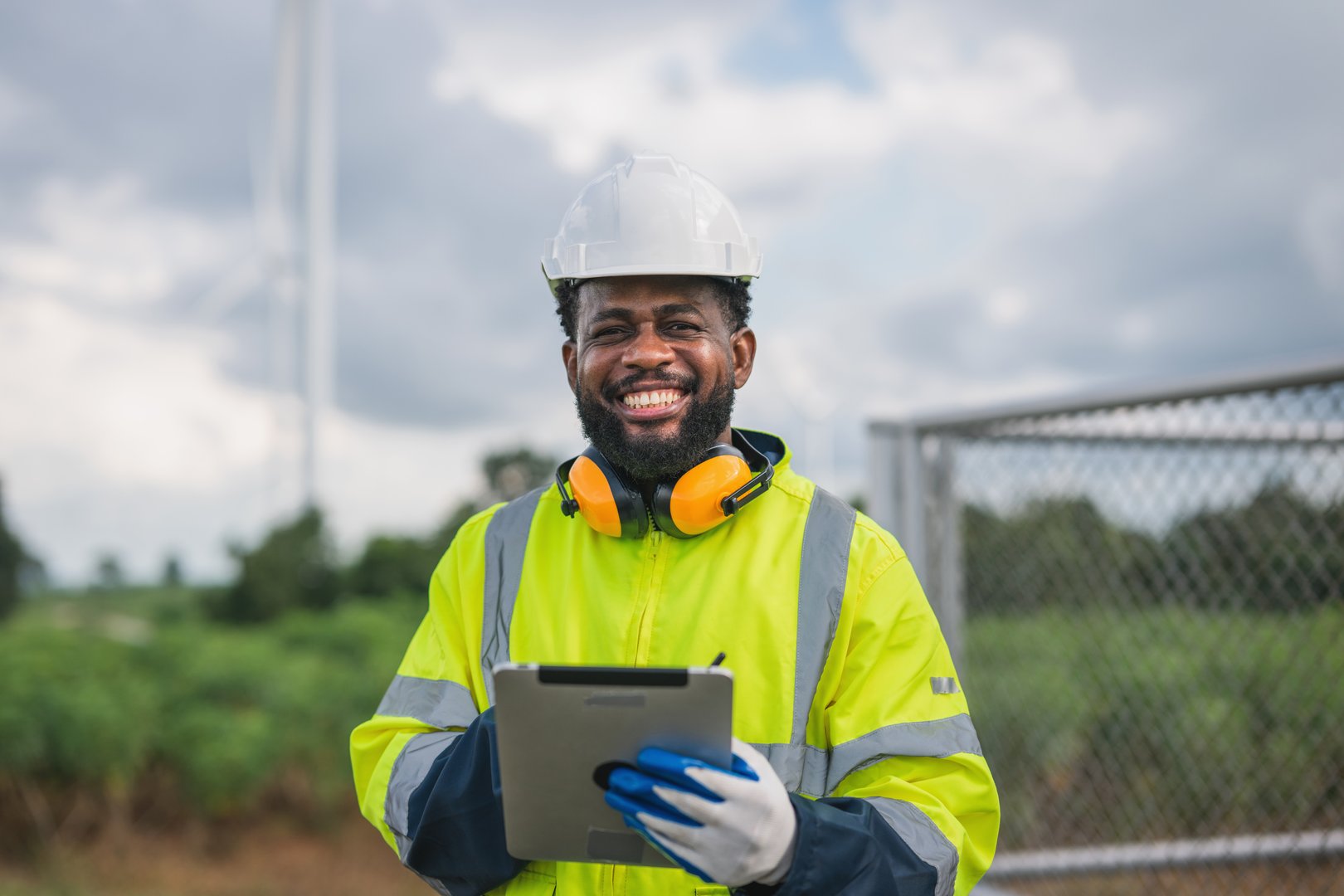 man in a yellow safety jacket is smiling and holding a tablet. He is wearing a hard hat and safety glasses