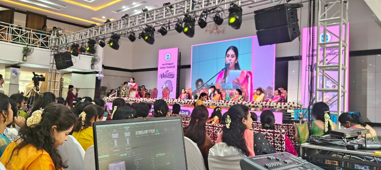 A woman speaks at a podium during an indoor event. Attendees sit facing the stage, with event lighting and a large screen visible.