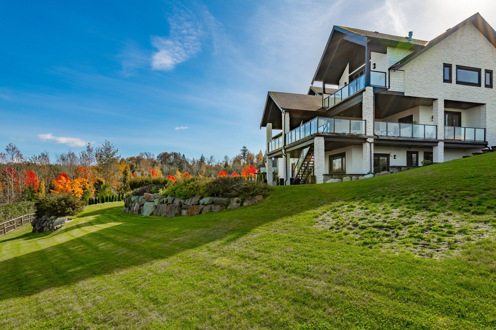 Elegant and stately home on a large lot on a sunny autumn fall day blue sky lush green grass colorful leaves on the trees the home is set against a deep blue sky and it is built with light colored stone and black wood trim black roof two large garages brick pillars and a wooden fence around a cobblestone driveway with a fountain in the centre and fully landscaped