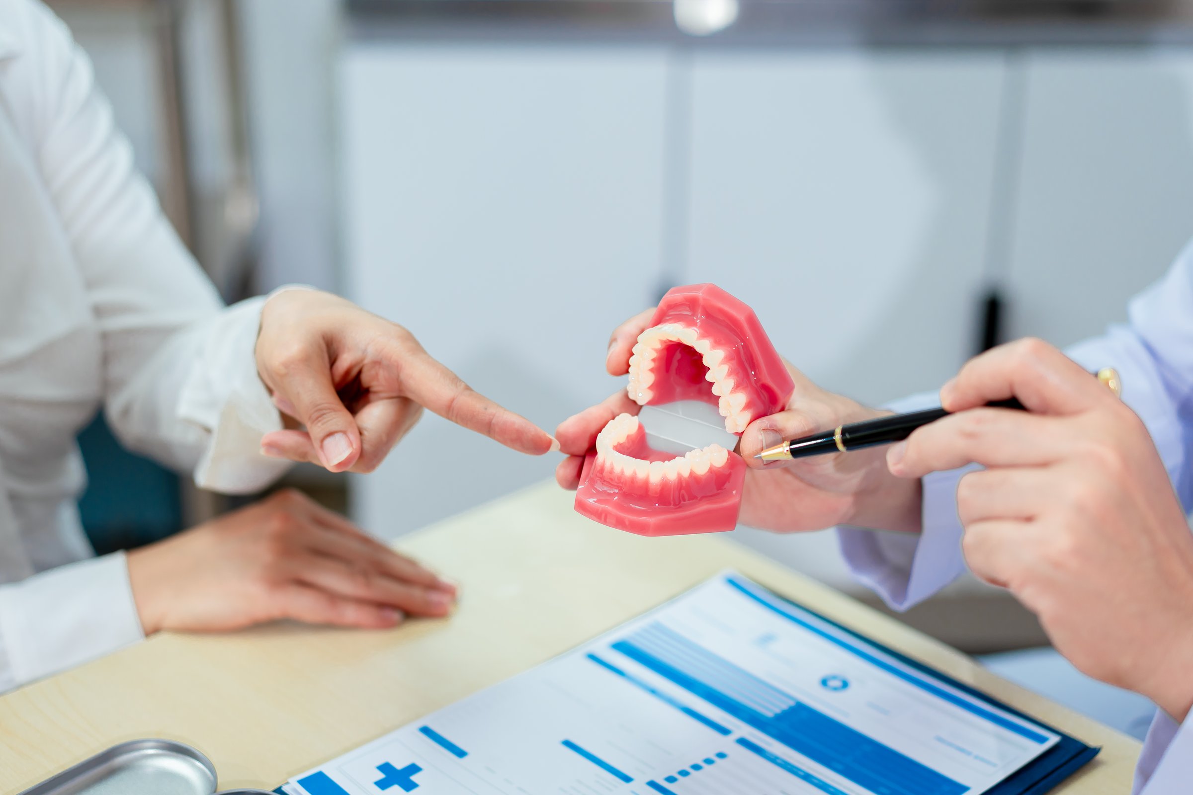An Asian male dentist explains dental health to a middle-aged Asian female patient while holding a dental model, demonstrating oral care and treatment procedures in a clinical consultation setting