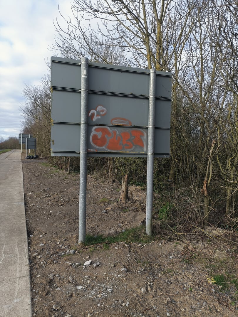A weathered roadside sign with graffiti, placed on a rural pathway next to bare trees and a patch of dirt and gravel.