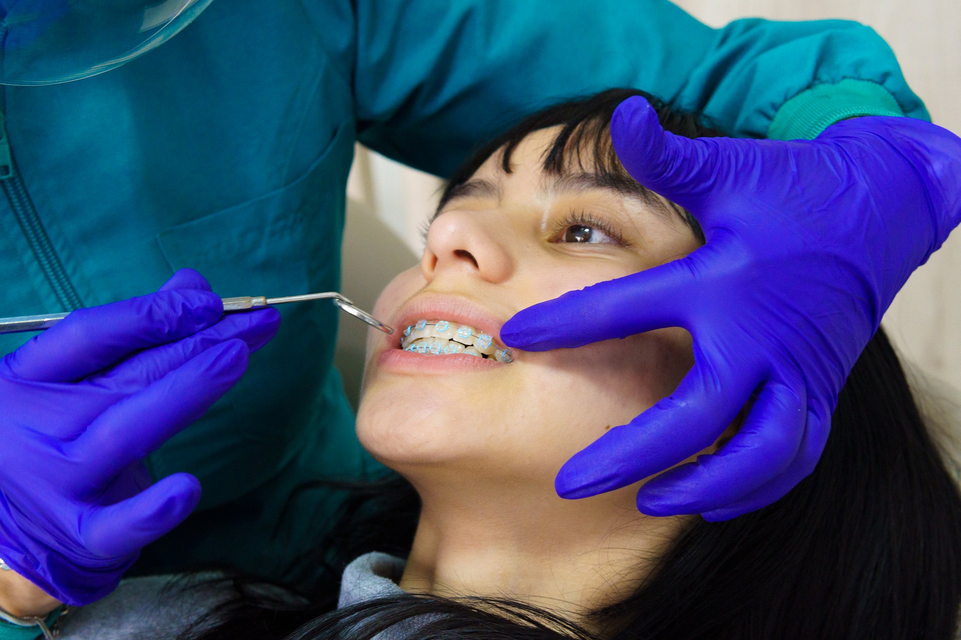A woman is getting her teeth cleaned by a dentist. The dentist is wearing blue gloves and holding a dental tool