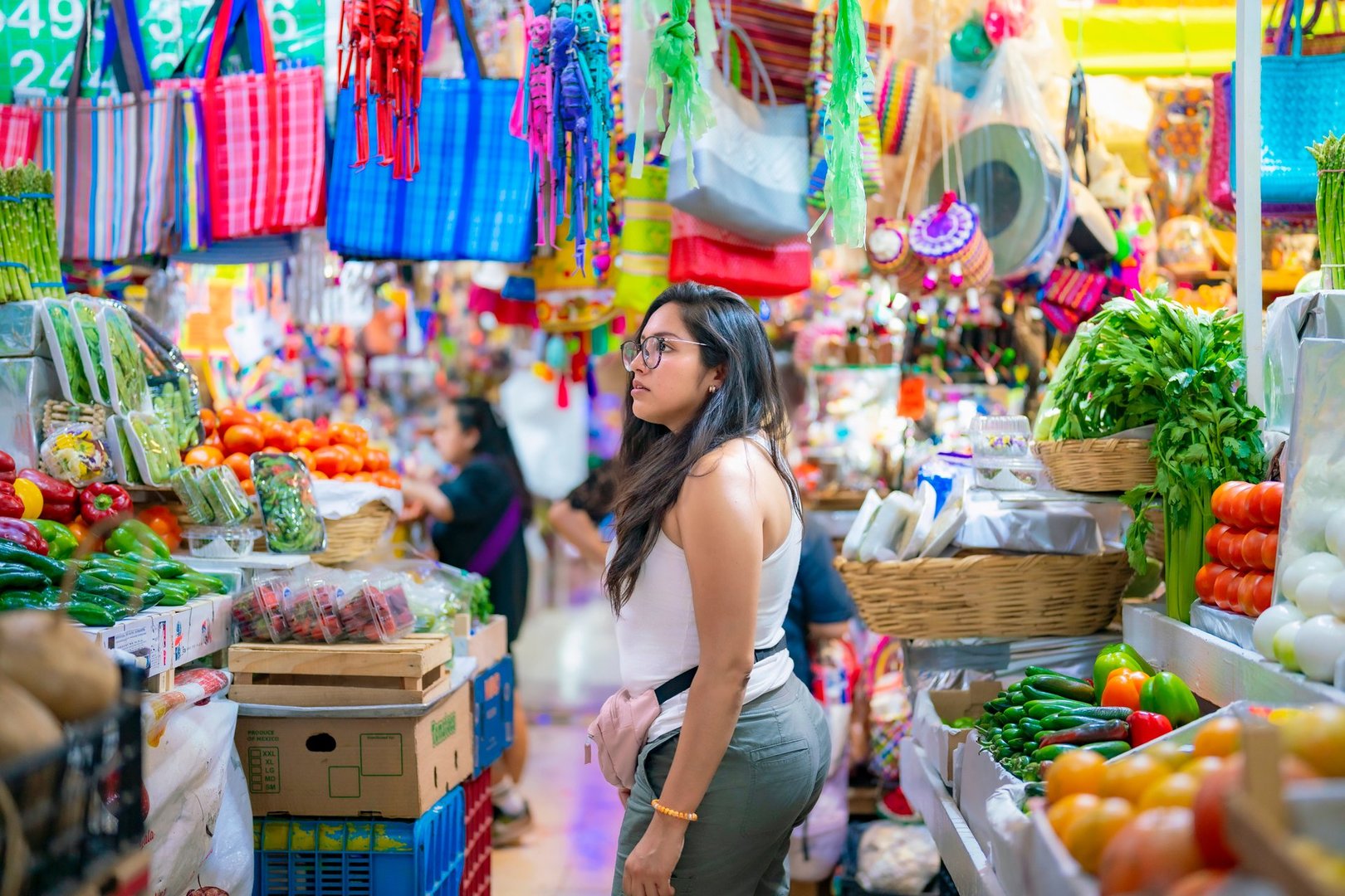 Young Latina woman exploring vibrant market stalls filled with fresh produce and colorful items in Mexico City, Mexico.