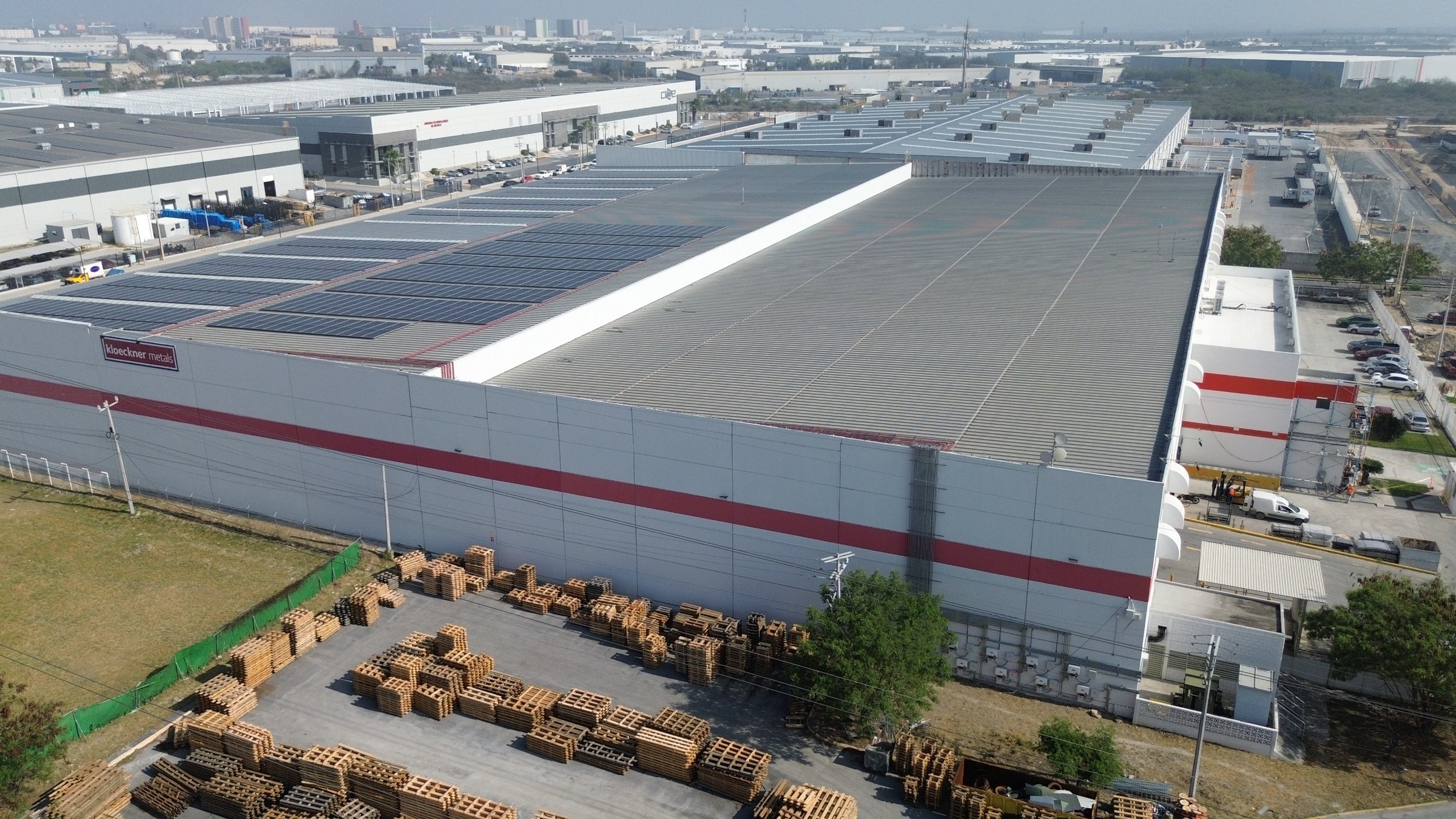 Aerial view of a large industrial warehouse with solar panels on the roof and numerous pallets stacked outside.