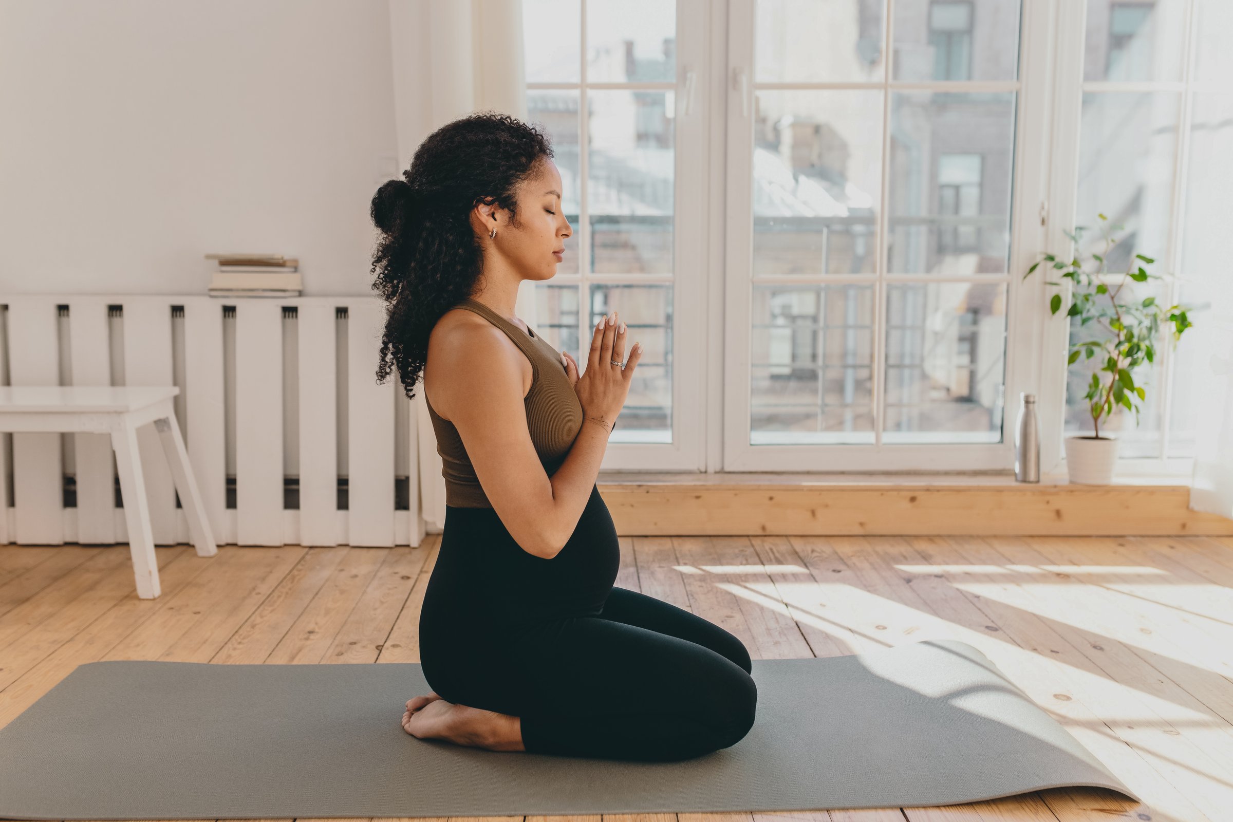Side view of gorgeous young pregnant african american woman standing on her knees in namaste posture sitting on mat on floor in living-room with closed eyes, meditating in beginning of yoga training