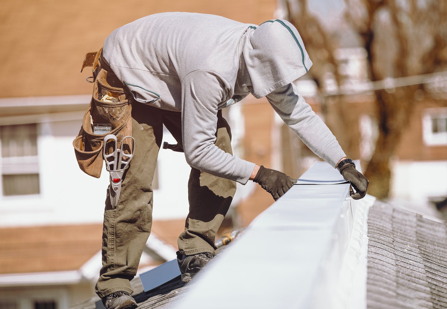 A roof repairman installs a metal ridge cap on top of a roof to seal and protect the roofline during maintenance. He is working with tools and safety gear under clear daylight, ensuring the roof's durability and weather resistance.