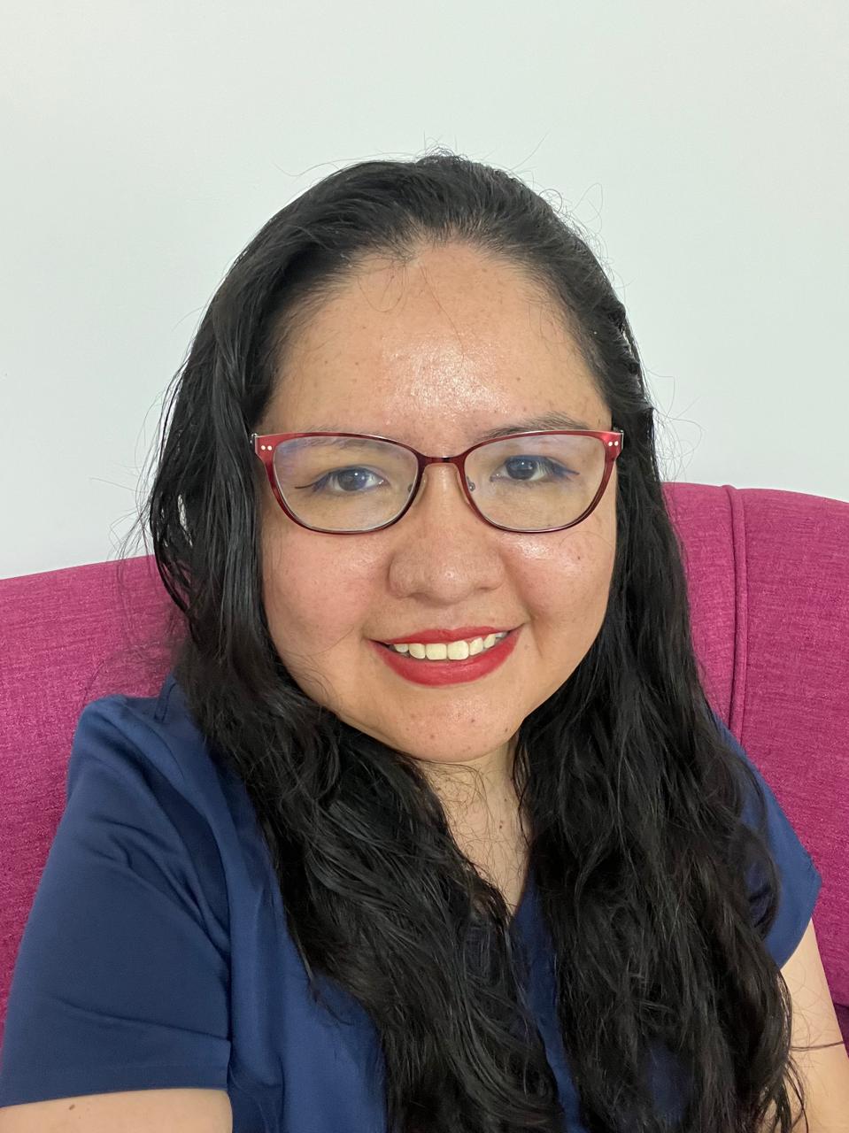 A woman with long dark hair and glasses smiles while sitting on a pink chair, wearing a blue shirt. The background is white.