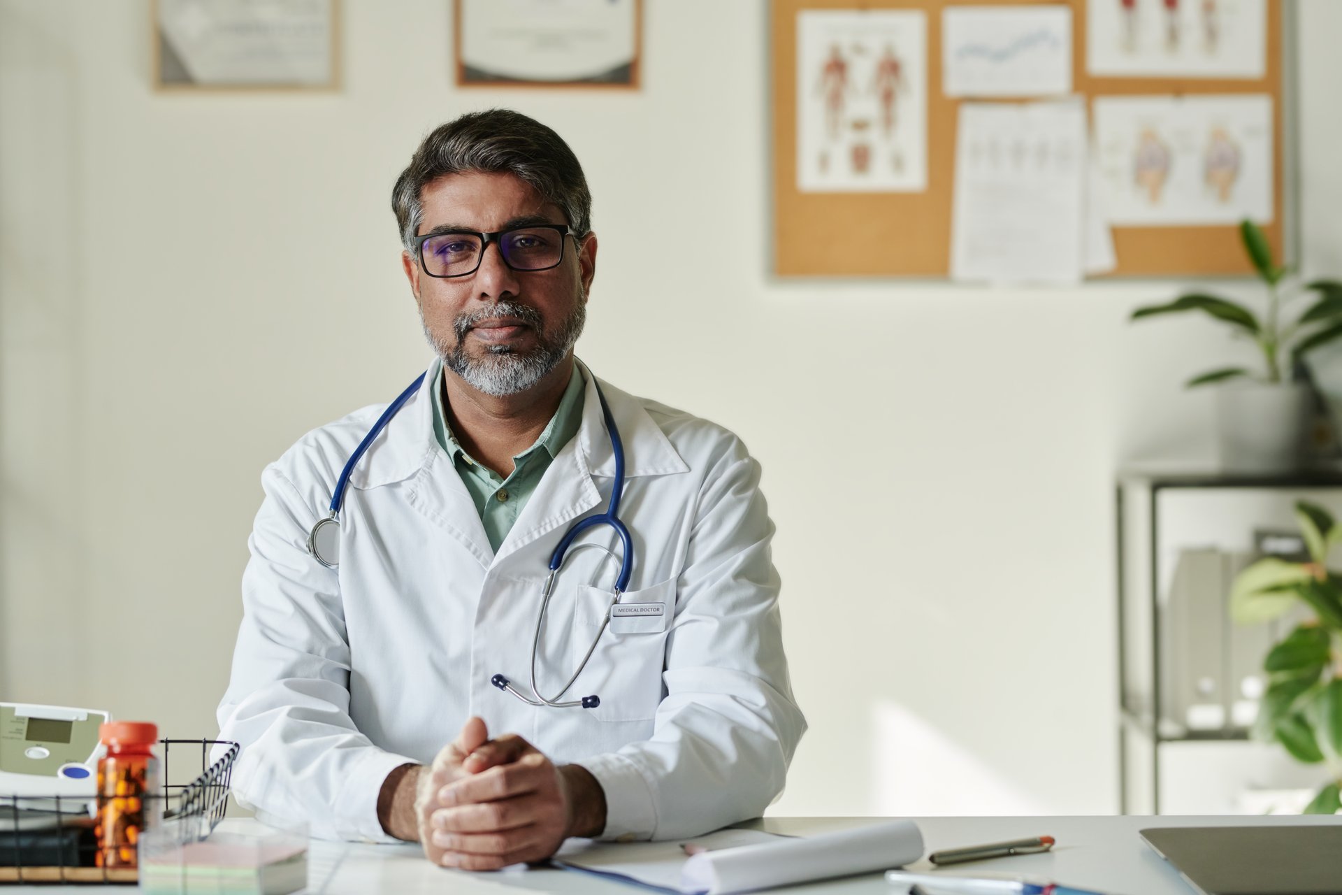 Serious mature male clinician with stethoscope looking at camera while sitting by workplace with medical document and other stuff