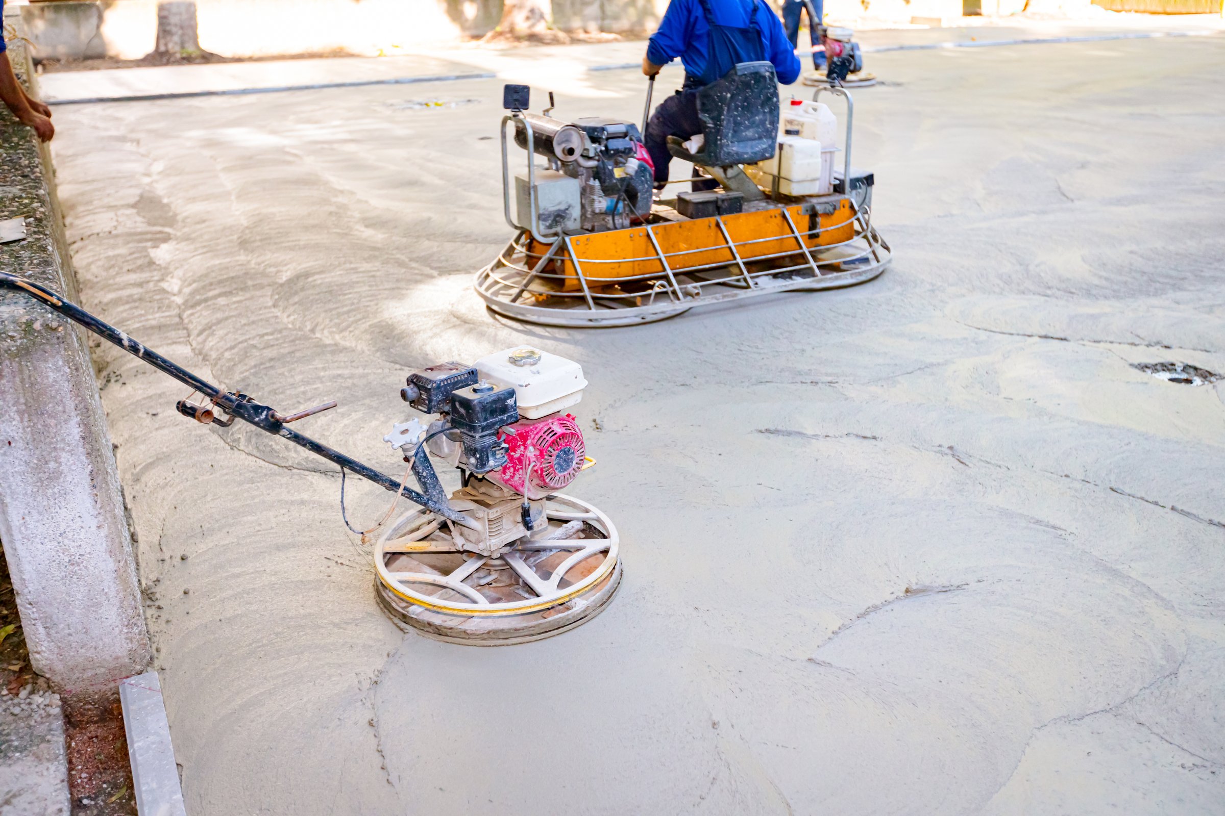 Two types of self-leveling power trowel machine with gas engine have wheels with pads, plate and he is driving it in circles smoothing surface on concrete slab.