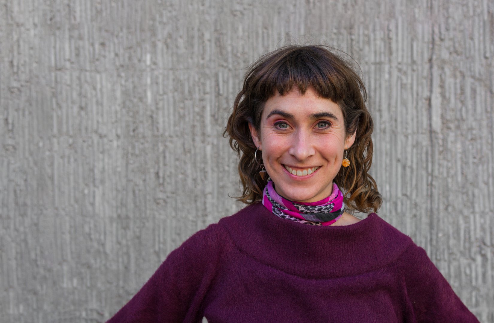 A simple style argentian woman smiling at camera ahead a gray wall | headshot | theatrical performer