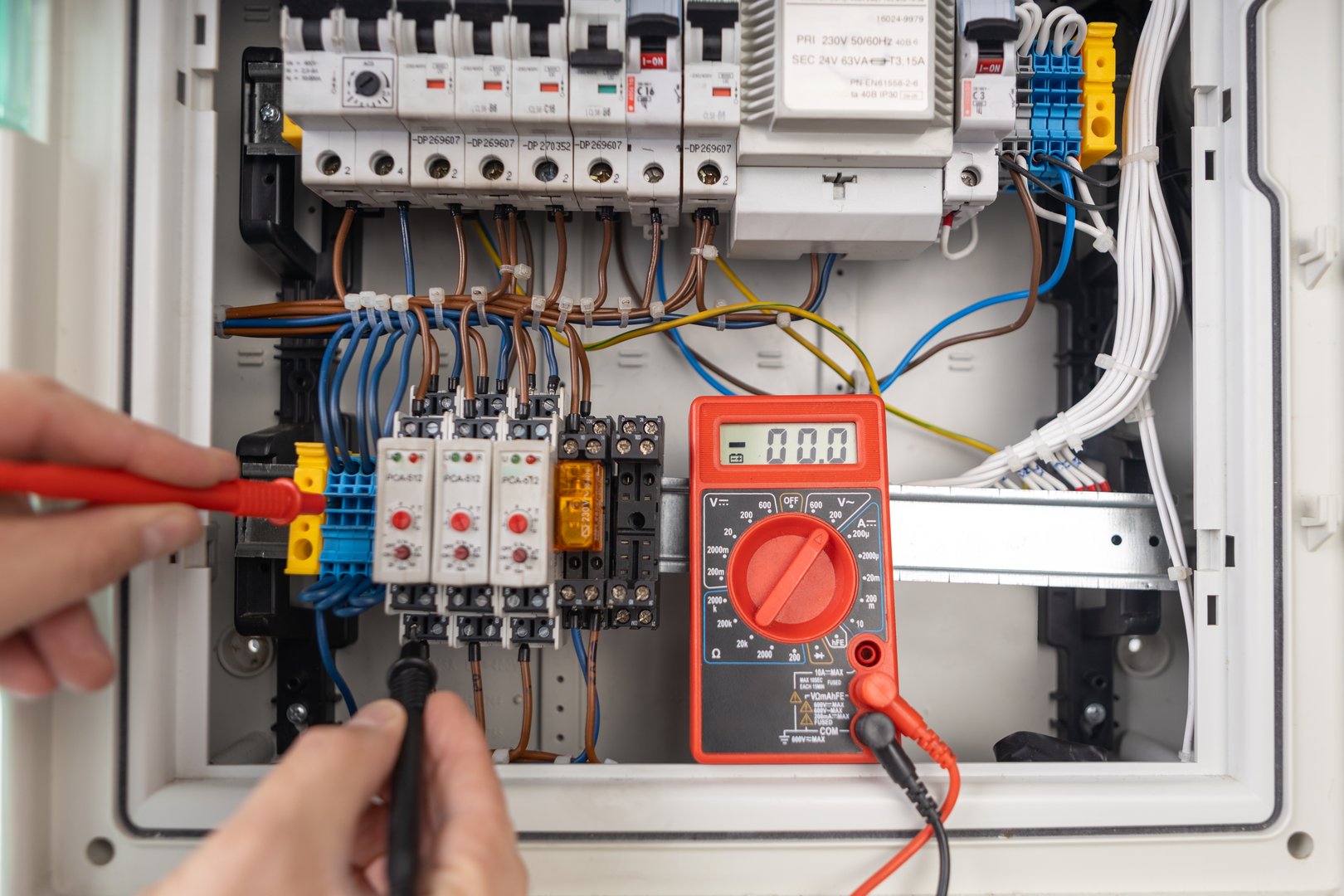 Hands using a digital multimeter to test an electrical circuit board with various connections and components