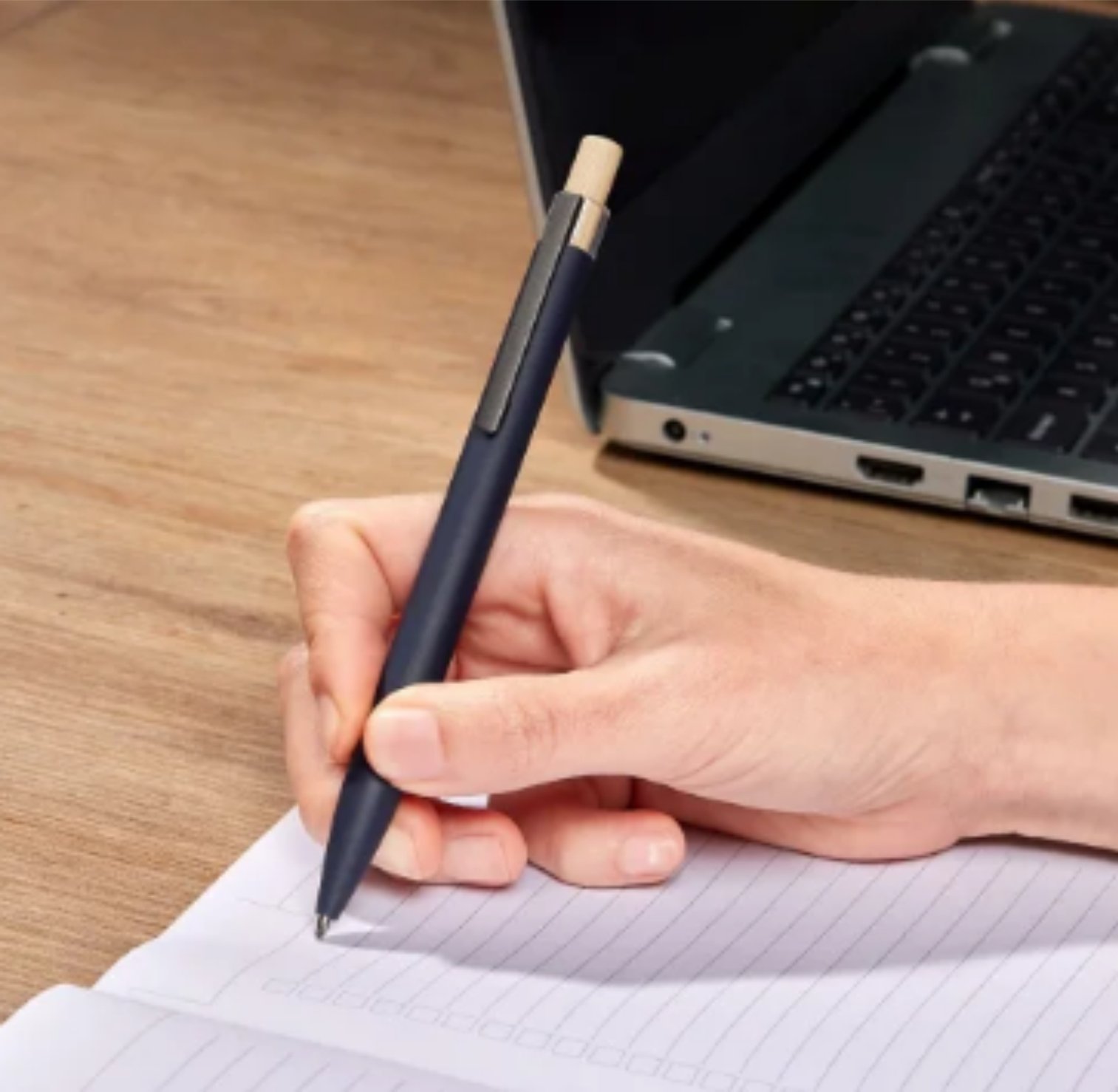 A hand writing with a pen on lined paper next to a laptop on a wooden desk.