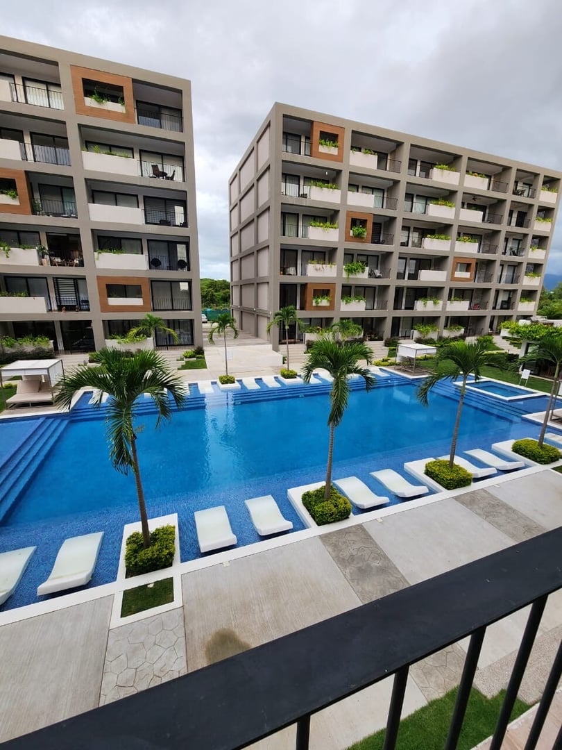 Modern apartment complex with two buildings, a large blue swimming pool, and surrounding palm trees.