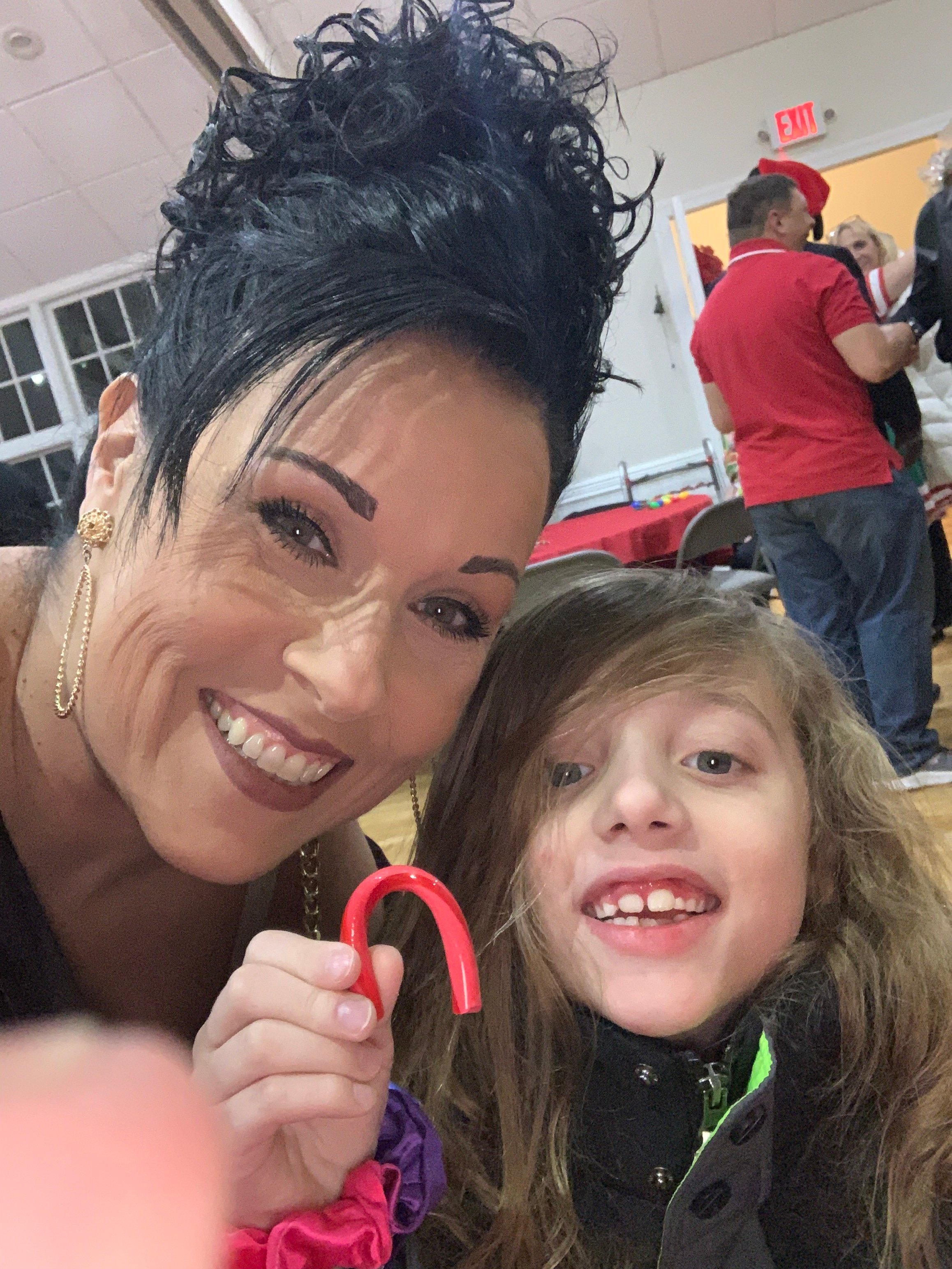 Woman and child smiling at an event, child holding a red candy cane. Indoors with people in the background.