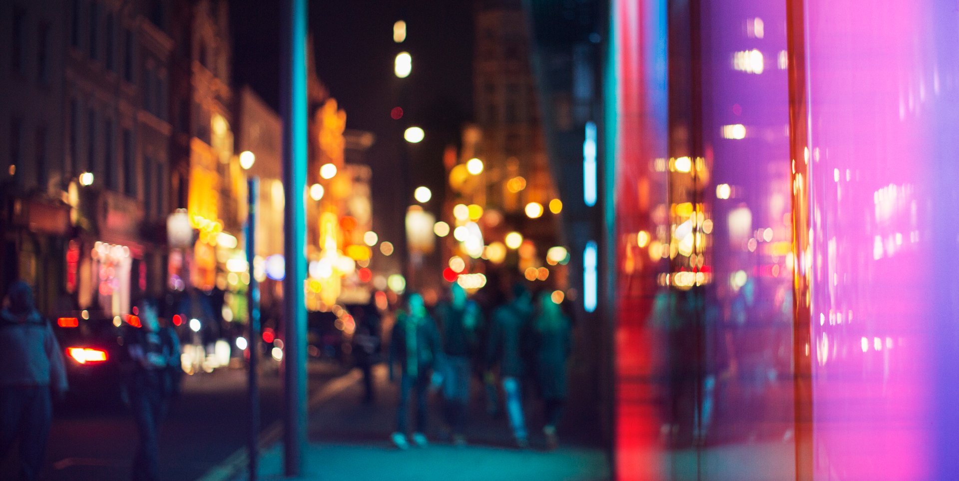 Blurry city nightlife scene with bright, colorful lights and silhouettes of people walking on a street.