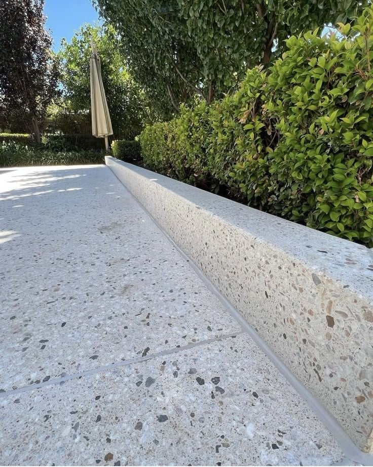 Concrete ledge with polished surface next to a hedge, umbrella, and trees under a clear blue sky.