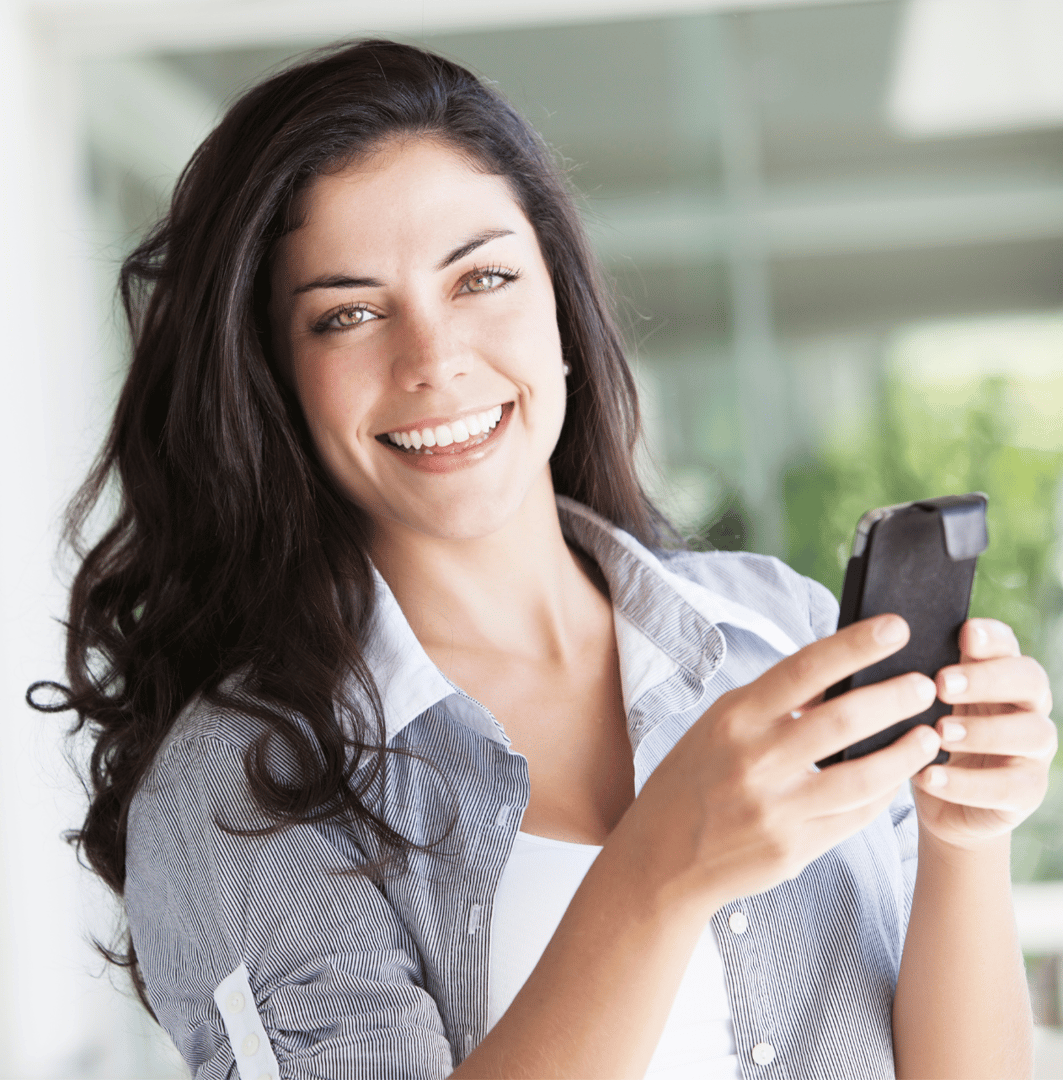 Smiling woman with long dark hair holds a smartphone in a bright indoor setting.