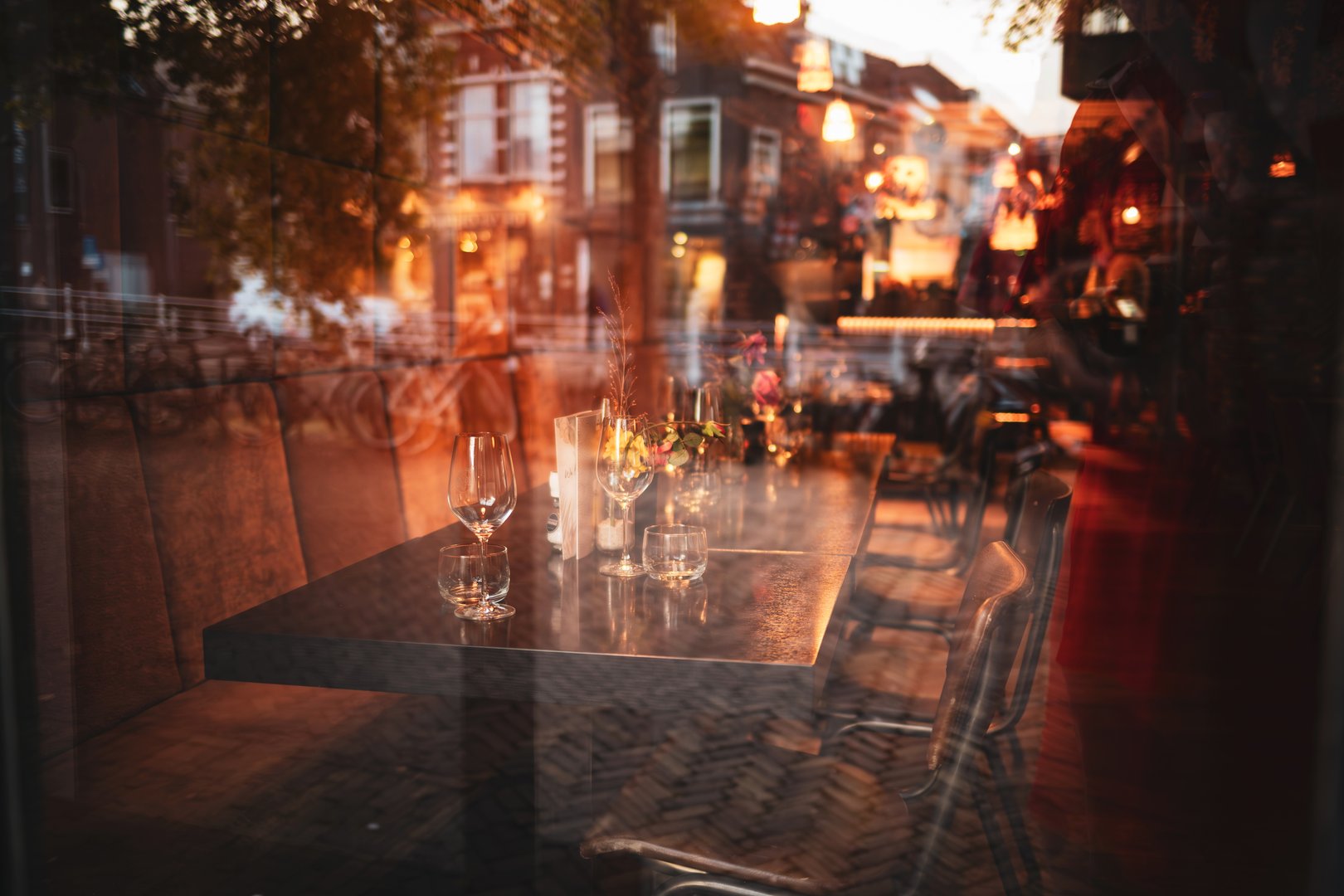 Empty restaurant table with wine glasses and flower arrangements as seen through the glass window from outside. Late evening light, shallow depth of field, window reflection of the street, no people