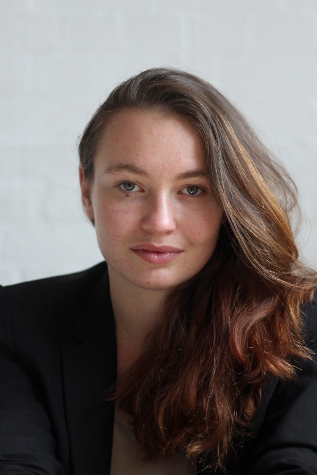 A person with long brown hair wearing a black blazer against a neutral background.