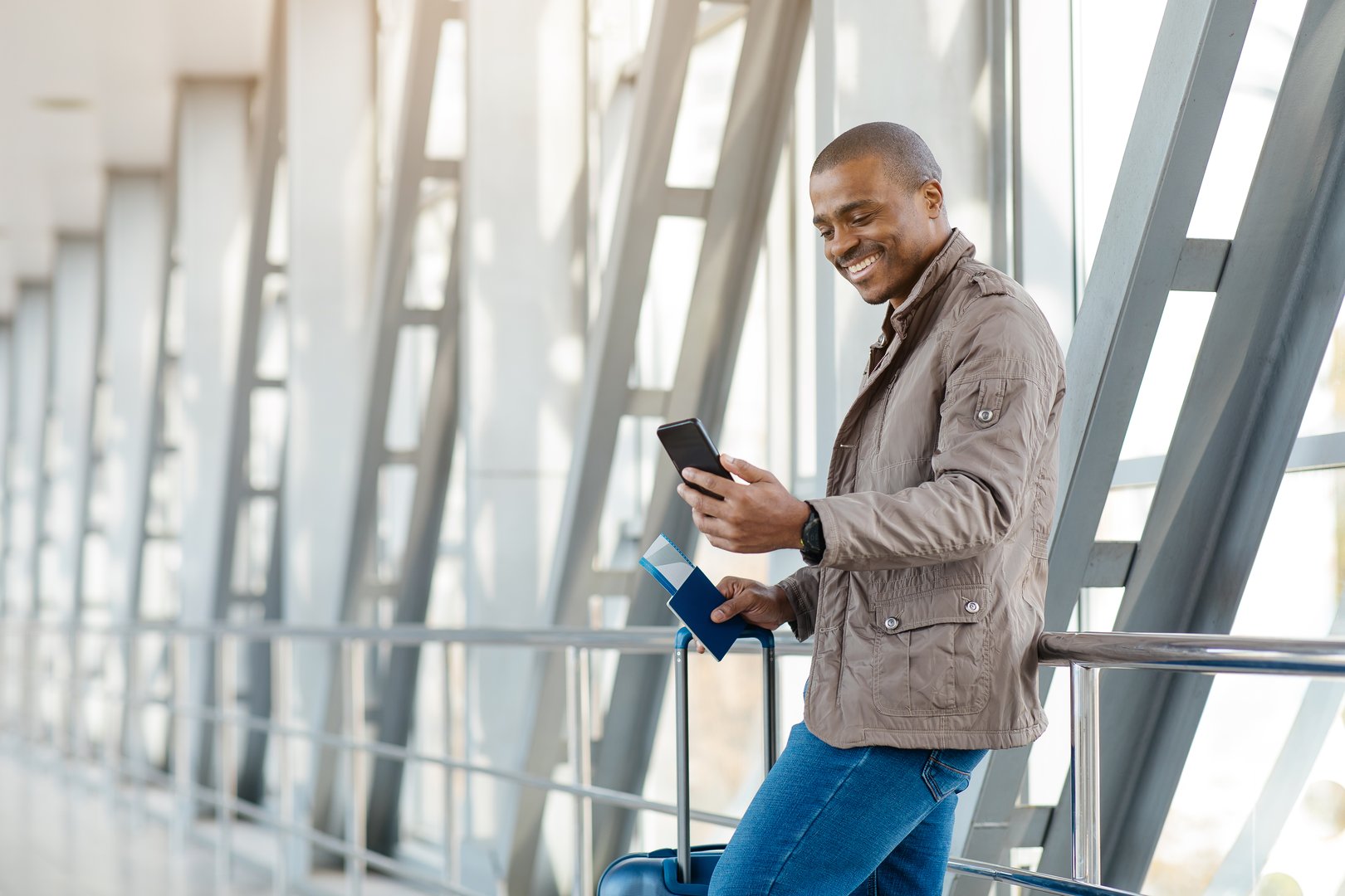 Smiling African Businessman Messaging On Smartphone While Waiting Flight At Airport, Handsome Young African American Male Standing With Suitcase At Terminal And Using Mobile Phone, Copy Space. High quality photo