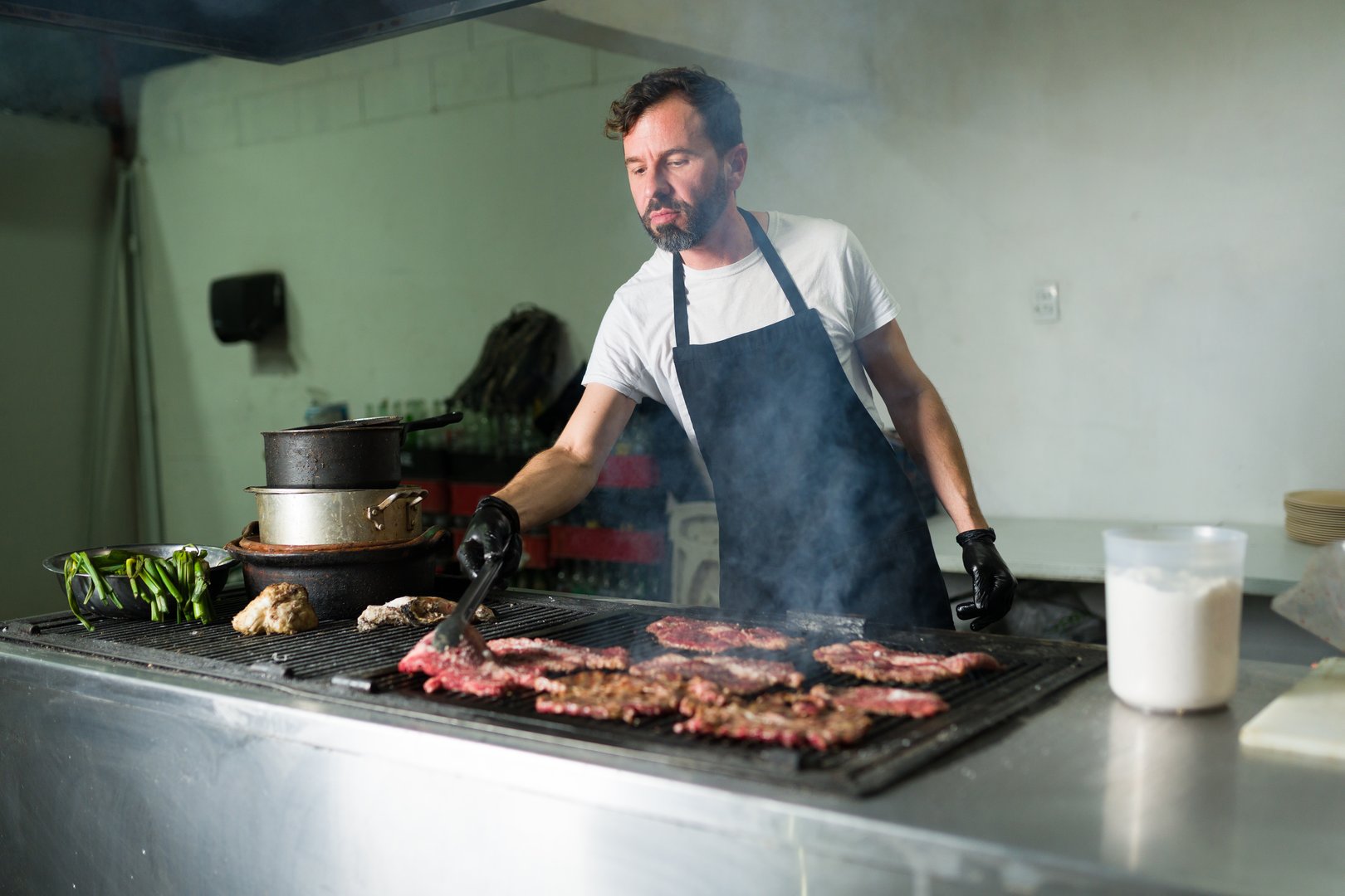 Latin cook in a restaurant kitchen grilling meat for tacos while wearing black gloves and an apron