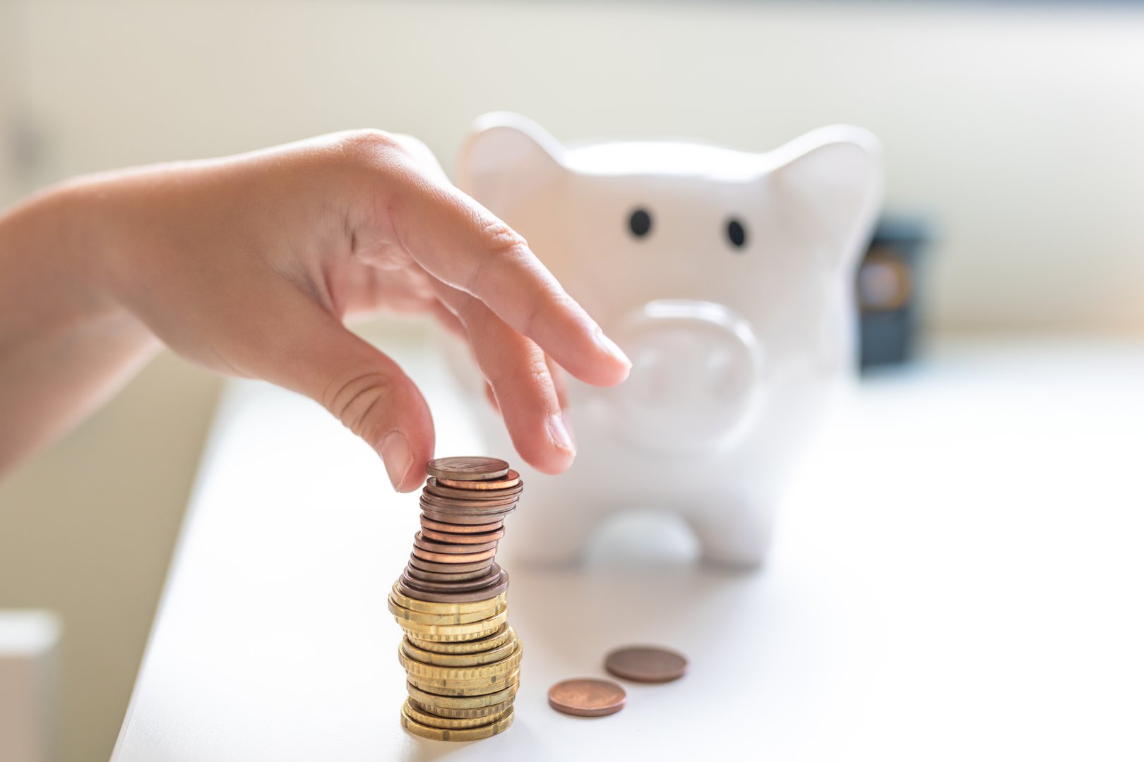 Close-up of a hand inserting a coin into a white piggy bank with scattered coins on a white table. Concept of saving money, finance, investment, and financial education.