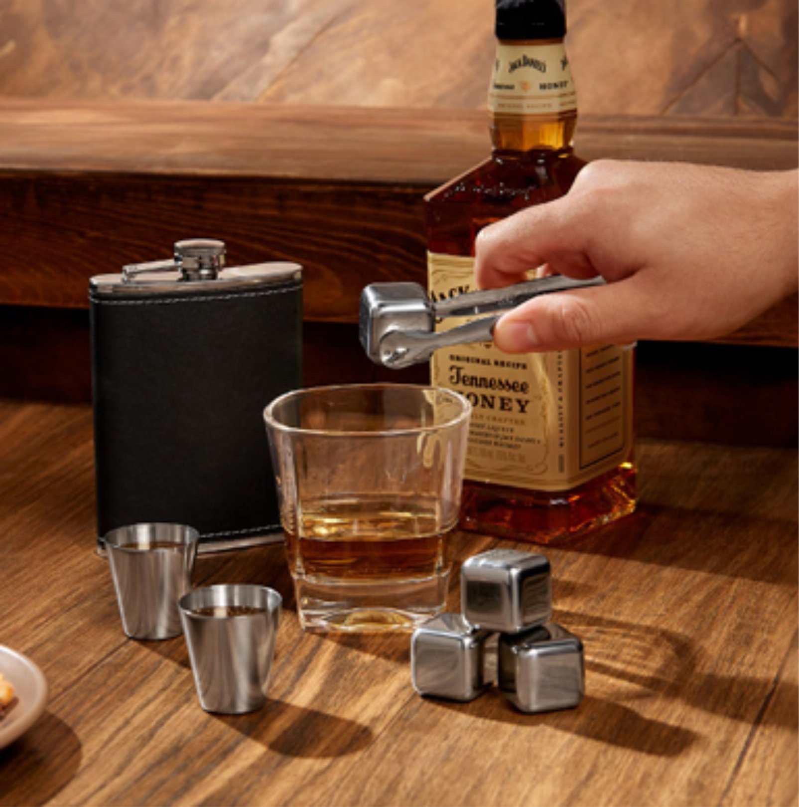 A glass of whiskey with stainless steel ice cubes, a flask, shot glasses, and a bottle of Tennessee Honey on a wooden table.