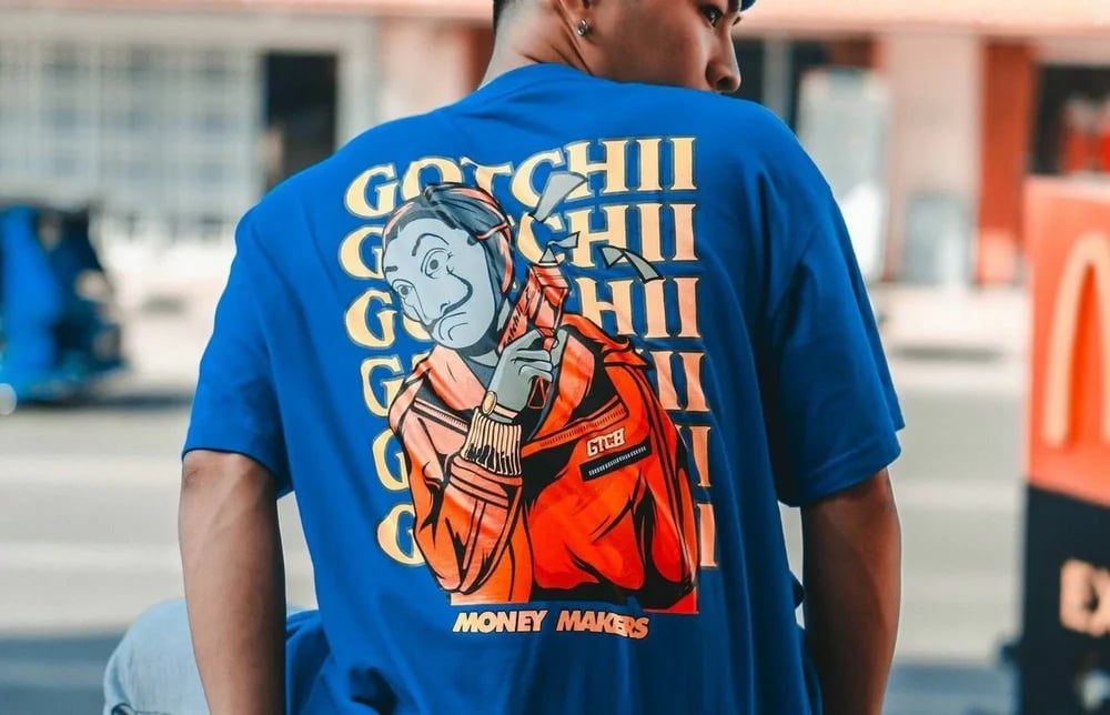 Person wearing a blue T-shirt featuring a large graphic of a character with a mask and text Gotchii Money Makers.