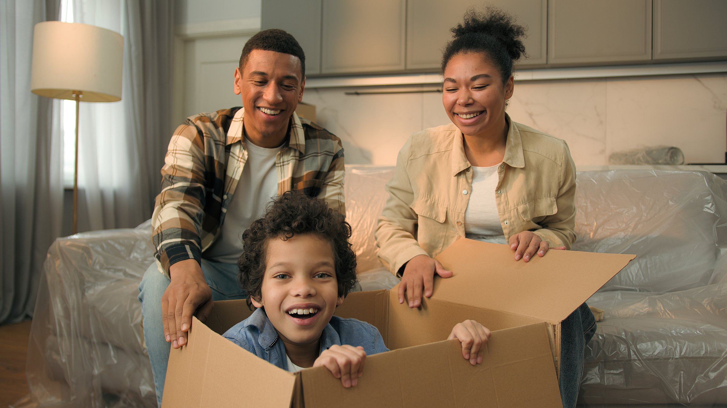 African American family moving in new home relocation happy parents unpacking parcel surprise funny joyful son little boy child jumping out of cardboard box playing game hide and seek kid having fun. High quality 4k footage