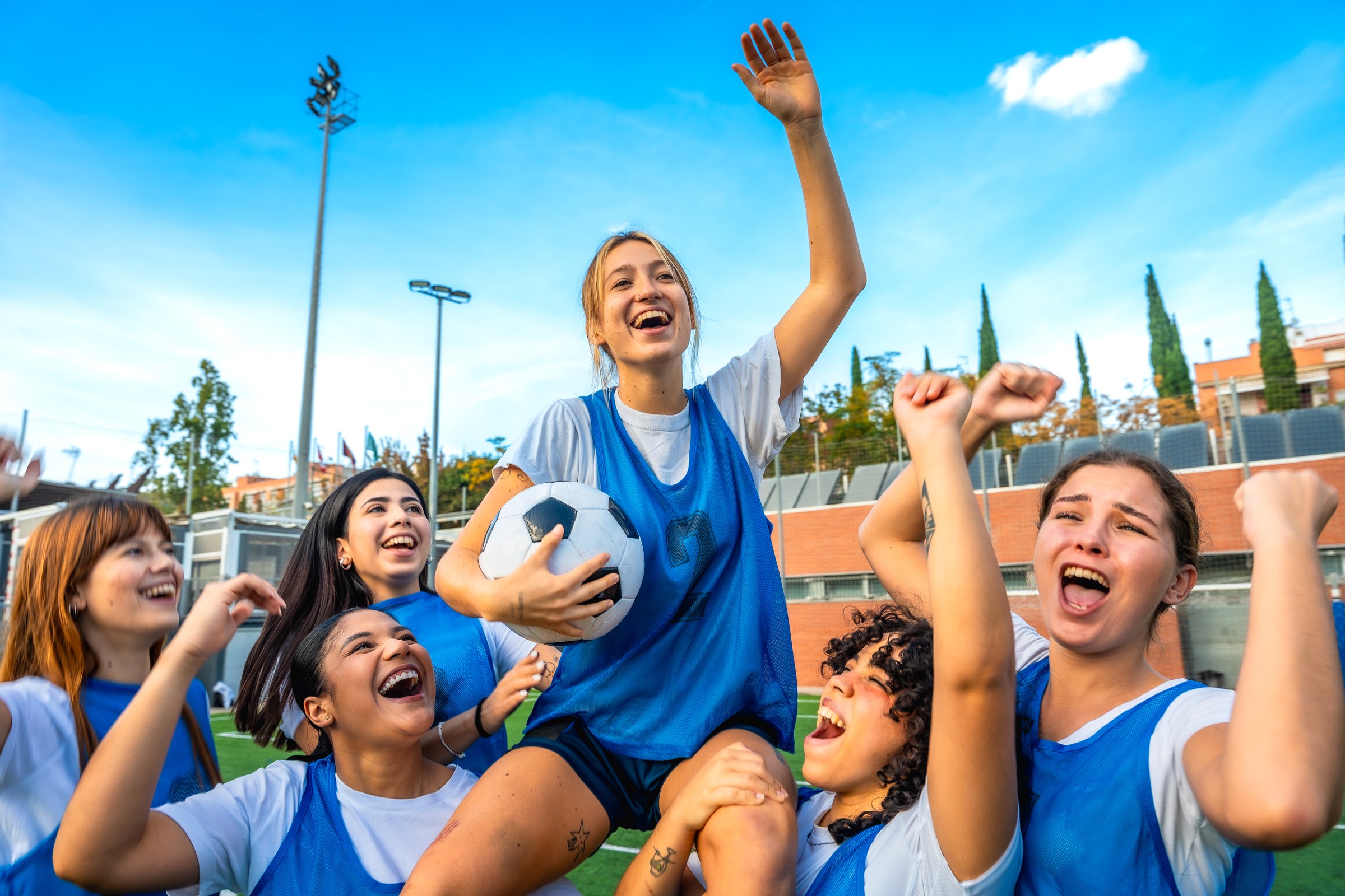 Female soccer teammates celebrate victory on a sunny field, lifting a teammate holding the ball high, smiling and cheering in a joyful display of teamwork and camaraderie