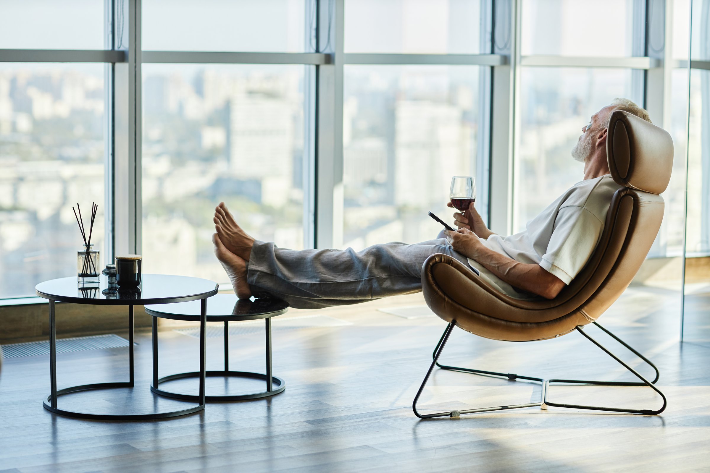 Side view of laid back successful mature man relaxing in comfy armchair with feet on footstool holding glass of red wine, while spending time in modern apartment with panoramic windows