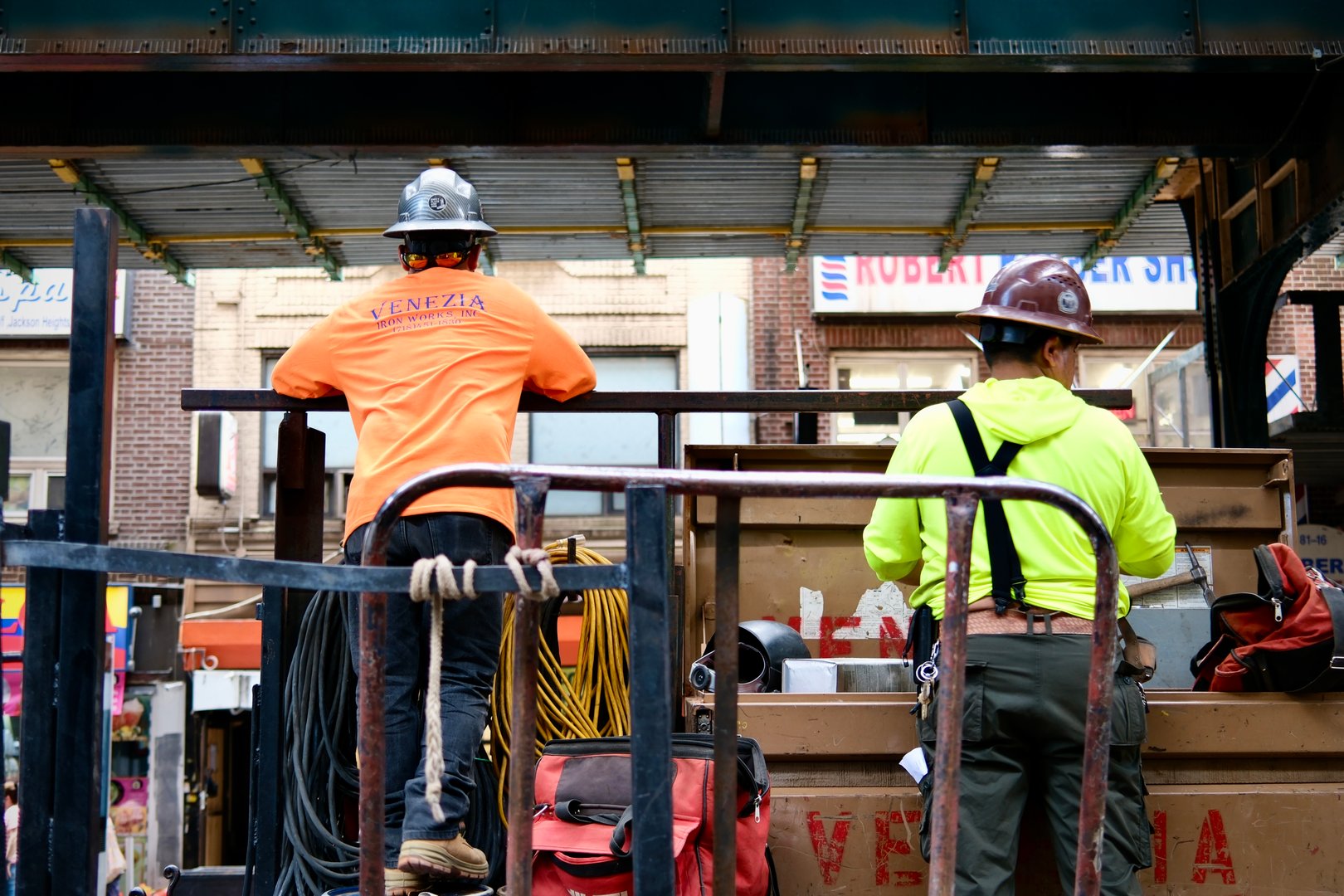 Queens, New York, USA – July 9, 2025: Two construction workers wearing hard hats and neon shirts load materials onto a truck beneath elevated subway tracks.