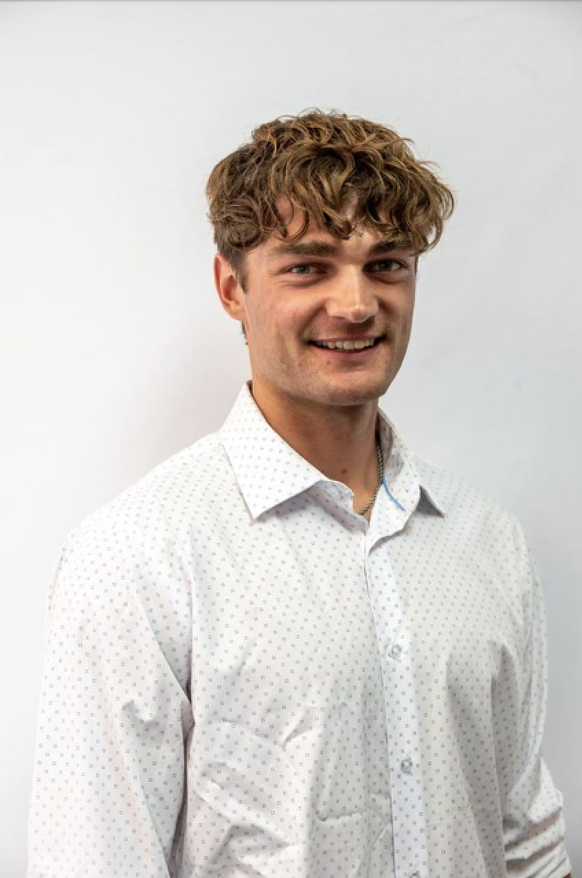 Smiling man with curly hair wearing a white button-up shirt stands against a plain background.