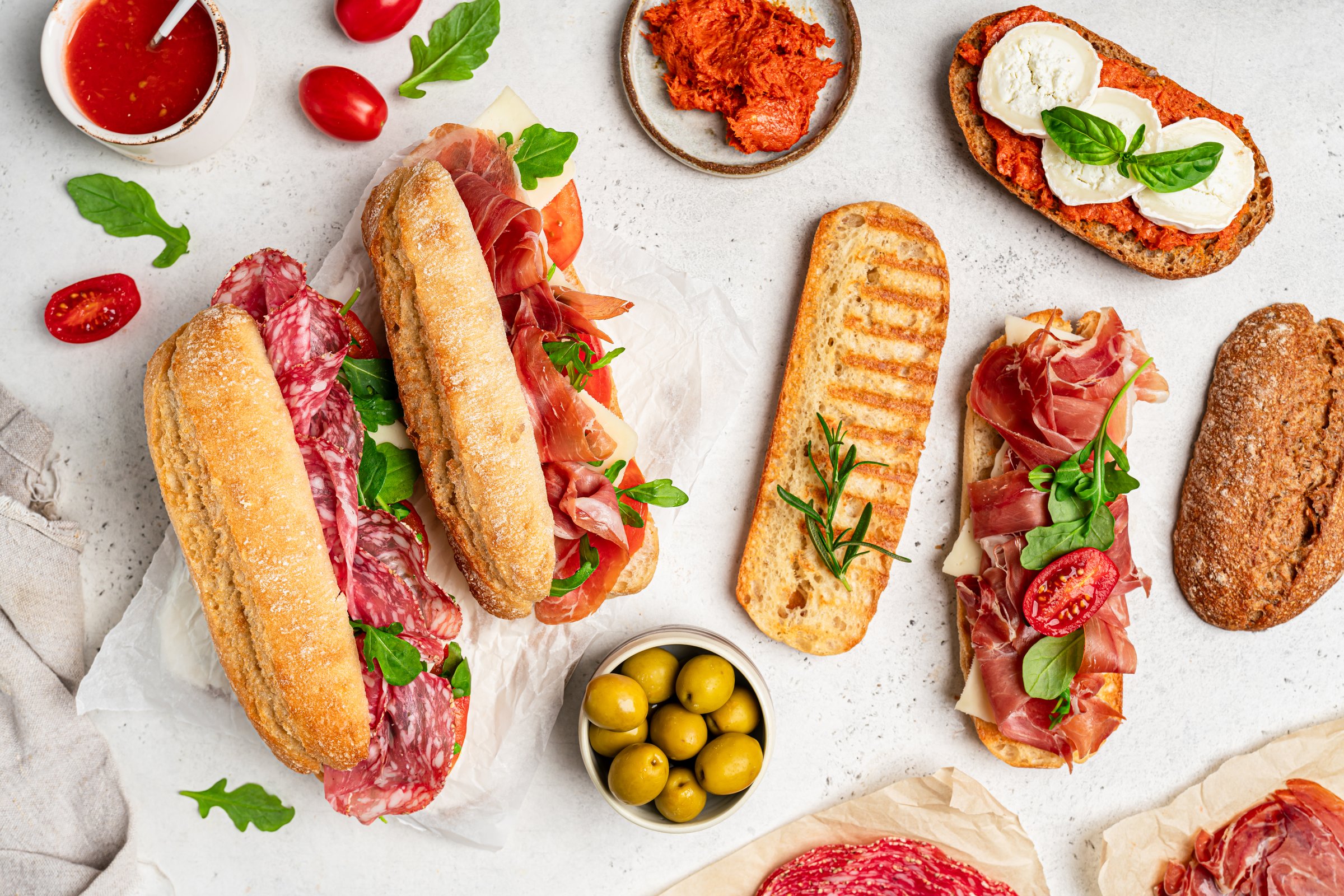 Assorted delicious baguette sandwiches filled with thinly sliced ham or salami and fresh green lettuce or arugula for breakfast arranged on white background, top view with copy space