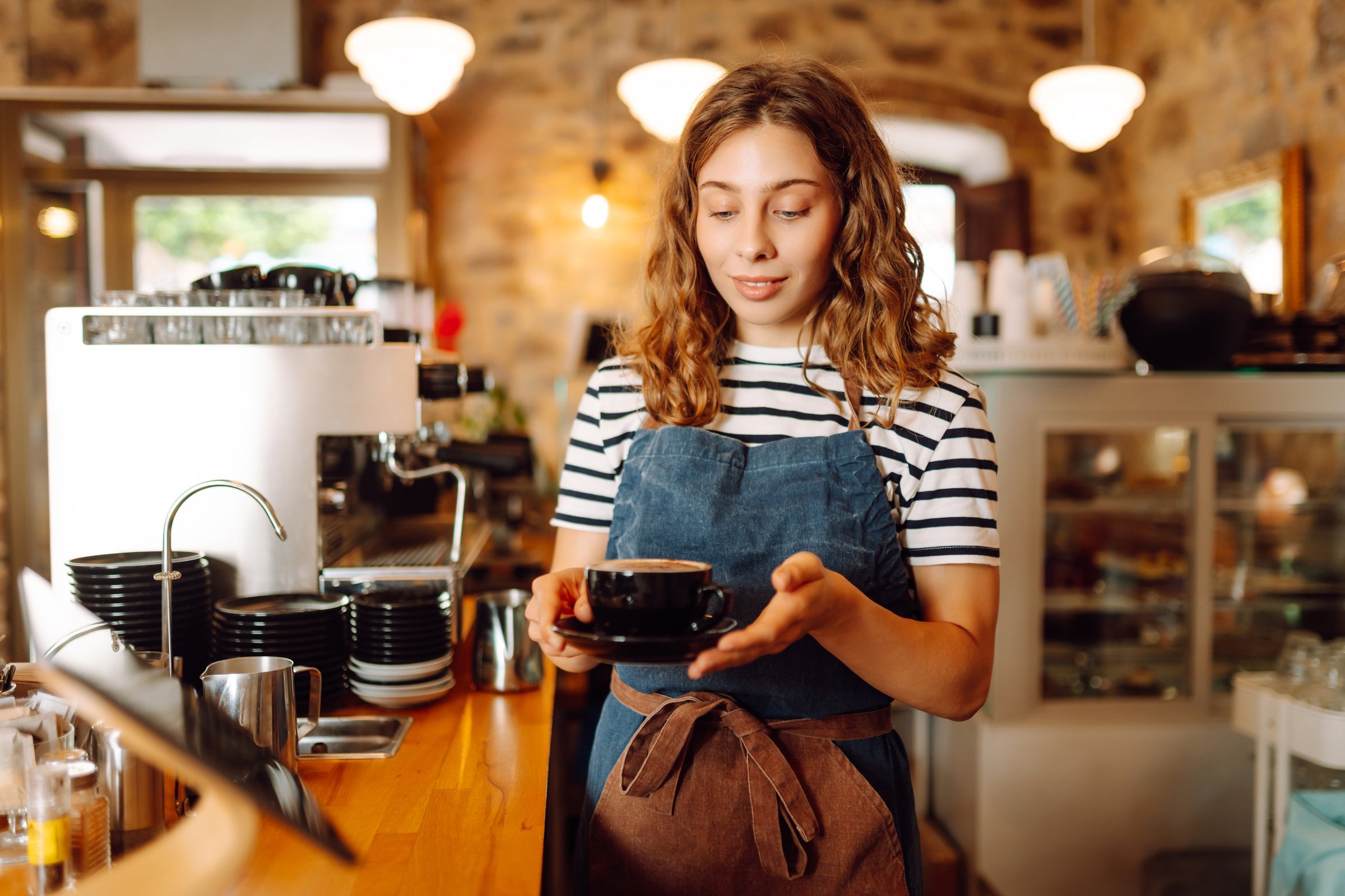 Happy female barista holding a cup of hot and aromatic coffee in her hands standing behind the bar. Young employee of the coffee shop gives coffee to a customer. Concept of drinks, small business.