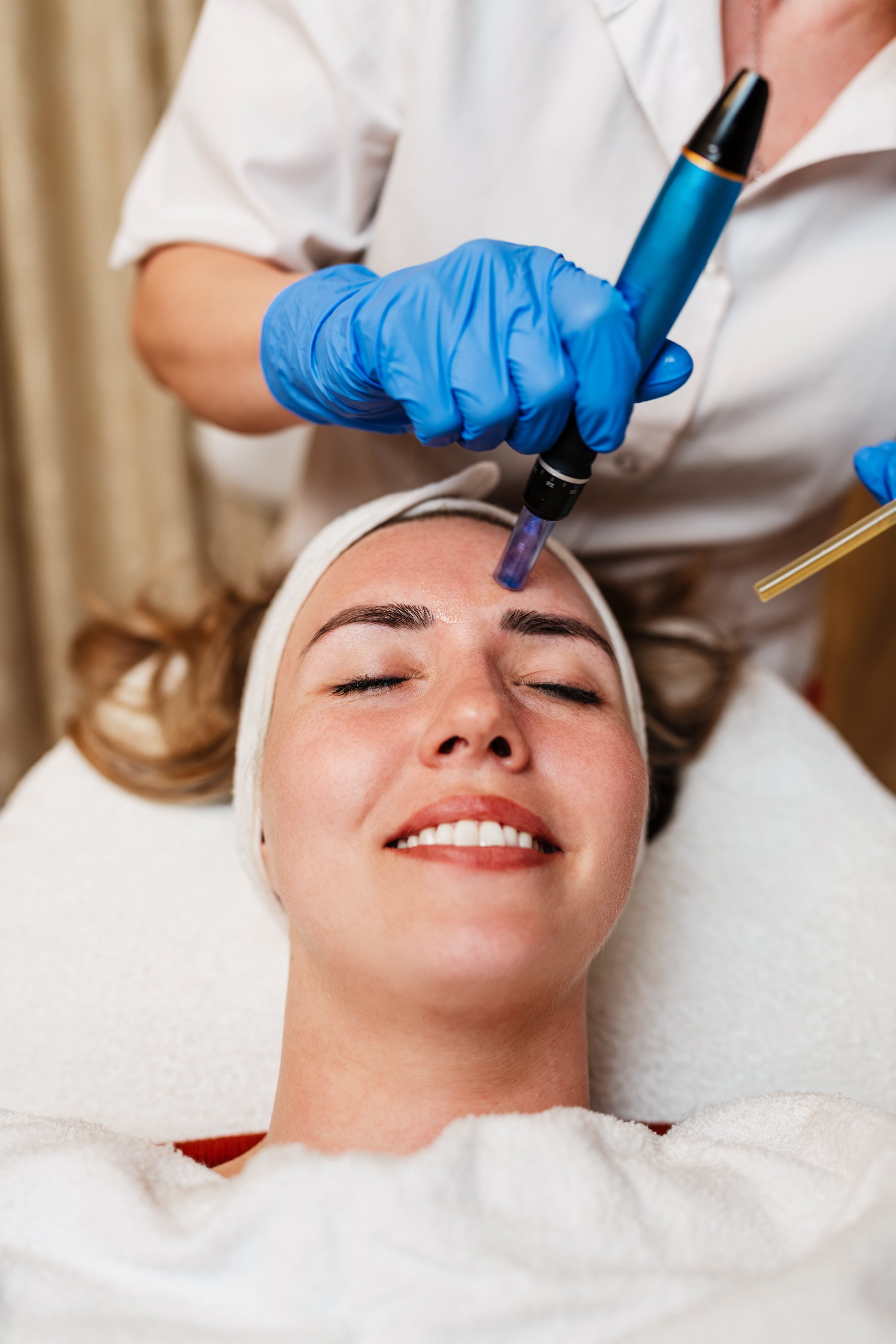 Beautician wearing blue gloves carefully applying serum to the forehead of a young woman during a microneedling treatment in a tranquil beauty spa, enhancing skin health and rejuvenation