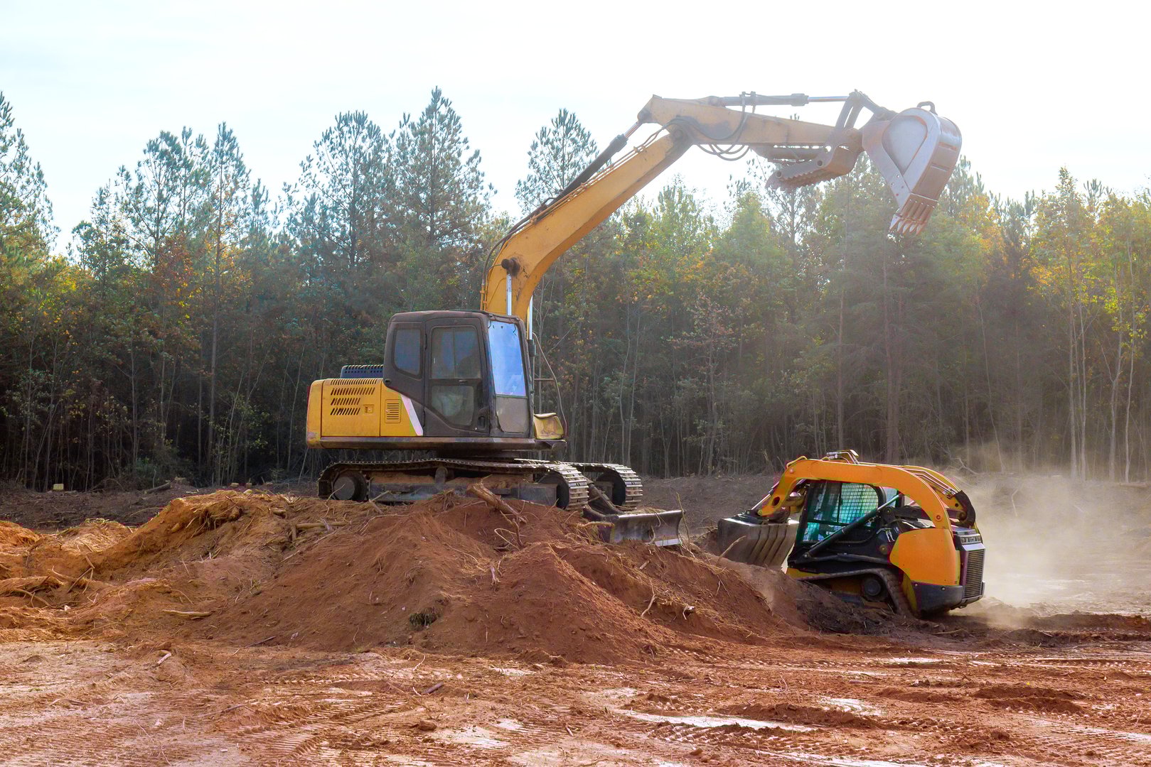 Heavy machinery with bulldozer, excavator performs land clearing on construction site, at dust rising amidst forest backdrop.