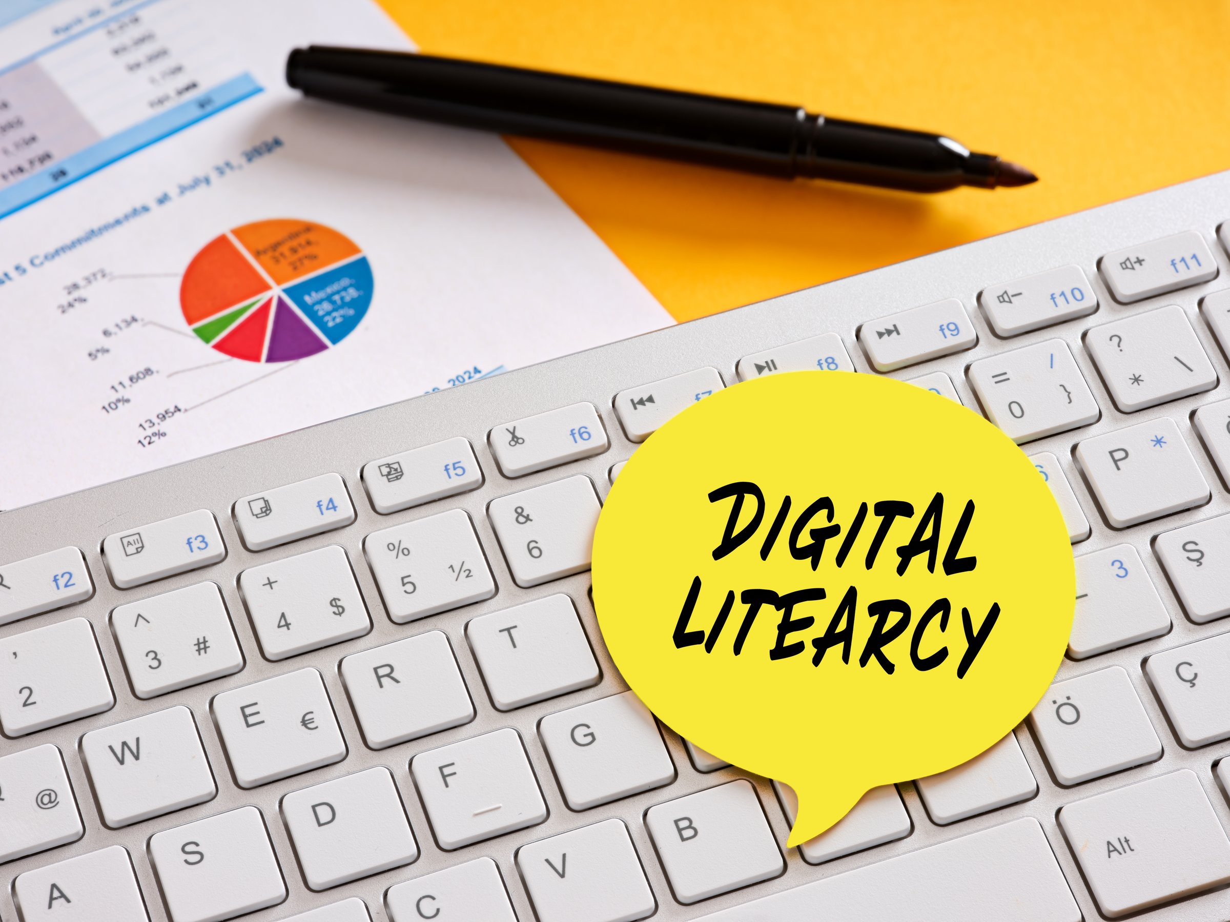 Yellow sticky note labeled digital literacy resting on a computer keyboard, surrounded by charts and a pen, representing online education