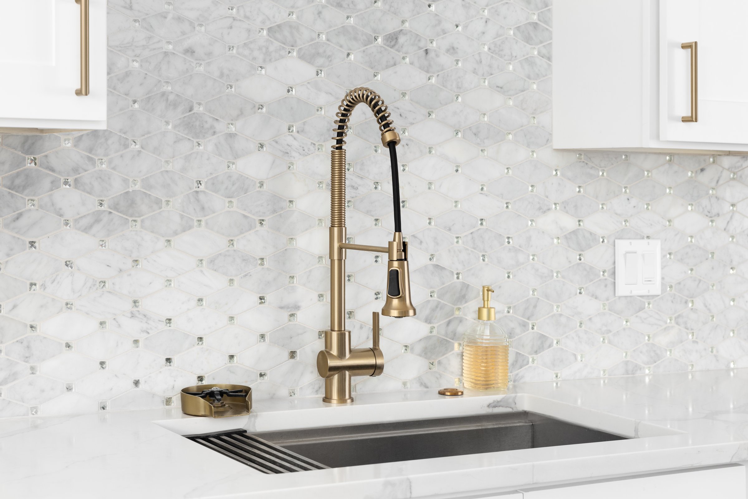 A kitchen with a gold faucet, white marble mosaic tile backsplash, white cabinets, and a white countertop.
