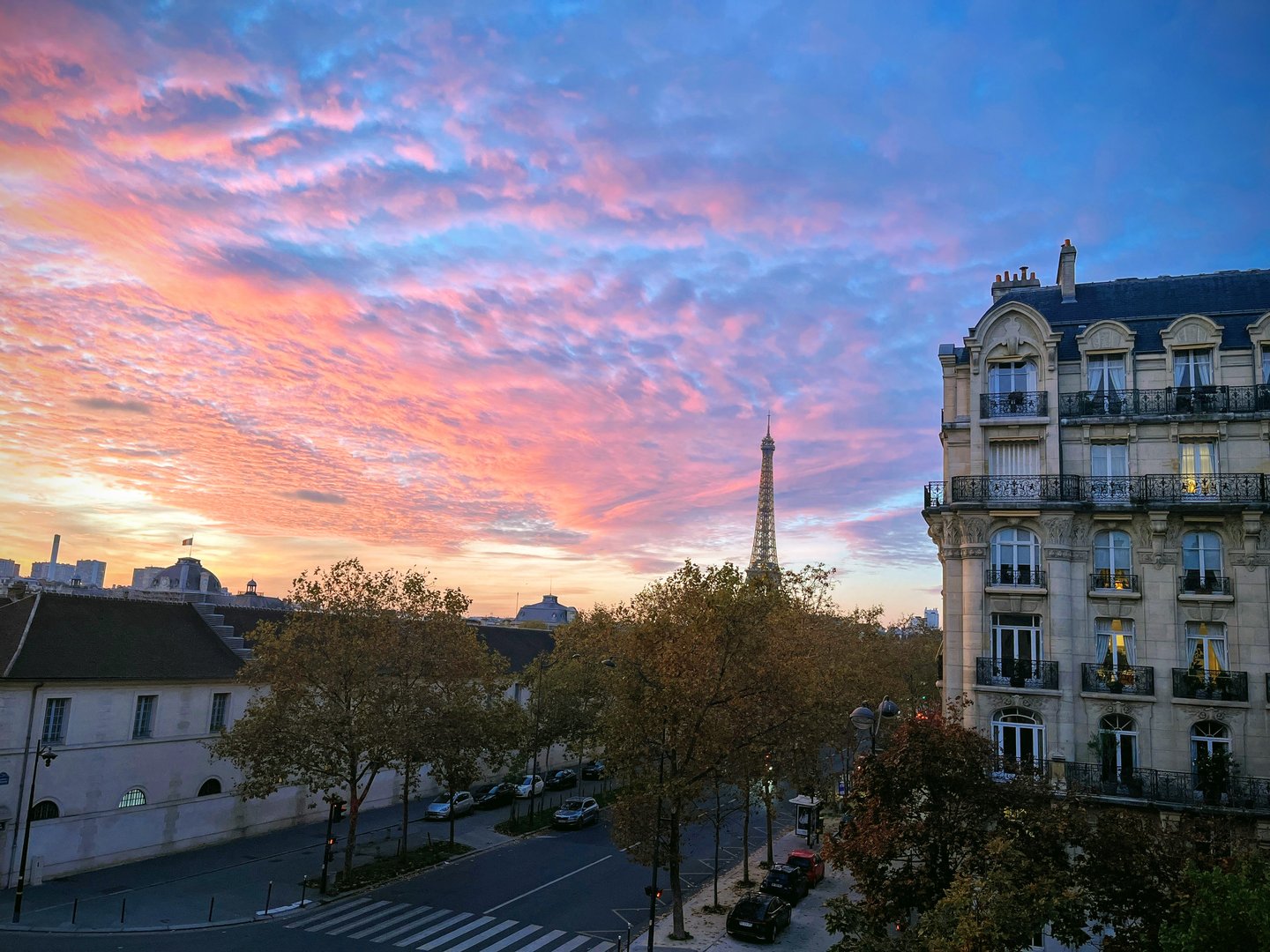 Parisian street view with Eiffel Tower in the background at sunset, vibrant clouds, and historic buildings.