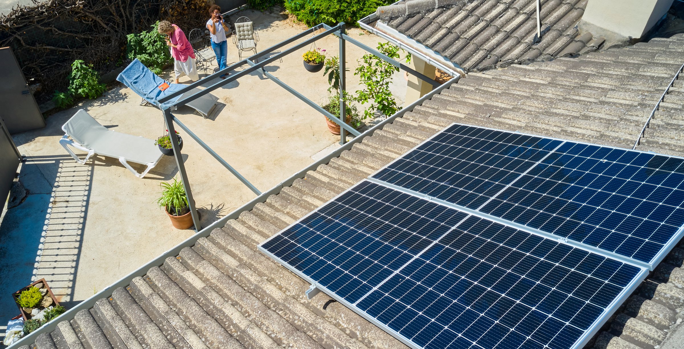 An aerial view of two women standing in a backyard, celebrating the installation of solar panels on the rooftop. The scene captures a moment of pride and environmental awareness, set in an eco-friendly home environment with greenery around.