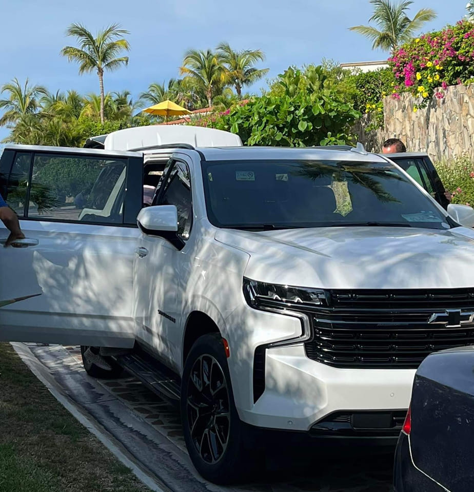 White SUV with open doors parked on a sunny day, surrounded by palm trees and colorful foliage.