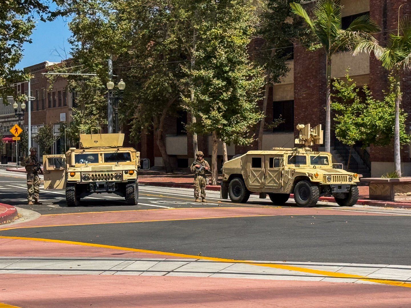 Editorial image of two Humvees and armed National Guard personnel stationed in downtown Santa Ana, California, during a daytime operation. The scene captures a moment of heightened security with soldiers standing alert near military vehicles on a city street surrounded by palm trees and historic brick buildings. This photo reflects themes of civil response, law enforcement, military readiness, and urban security measures in the United States.