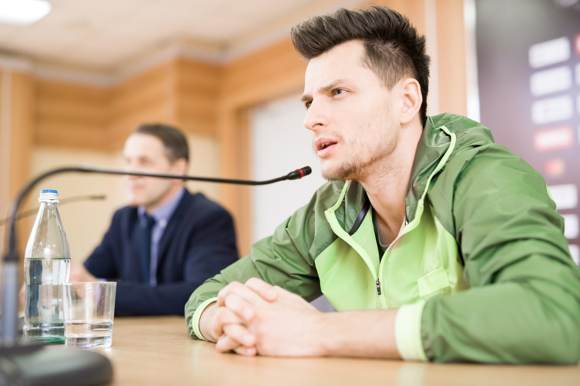 Concentrated young sportsman wearing track jacket sitting in front of microphone and listening to journalist while participating in press conference after come out on top
