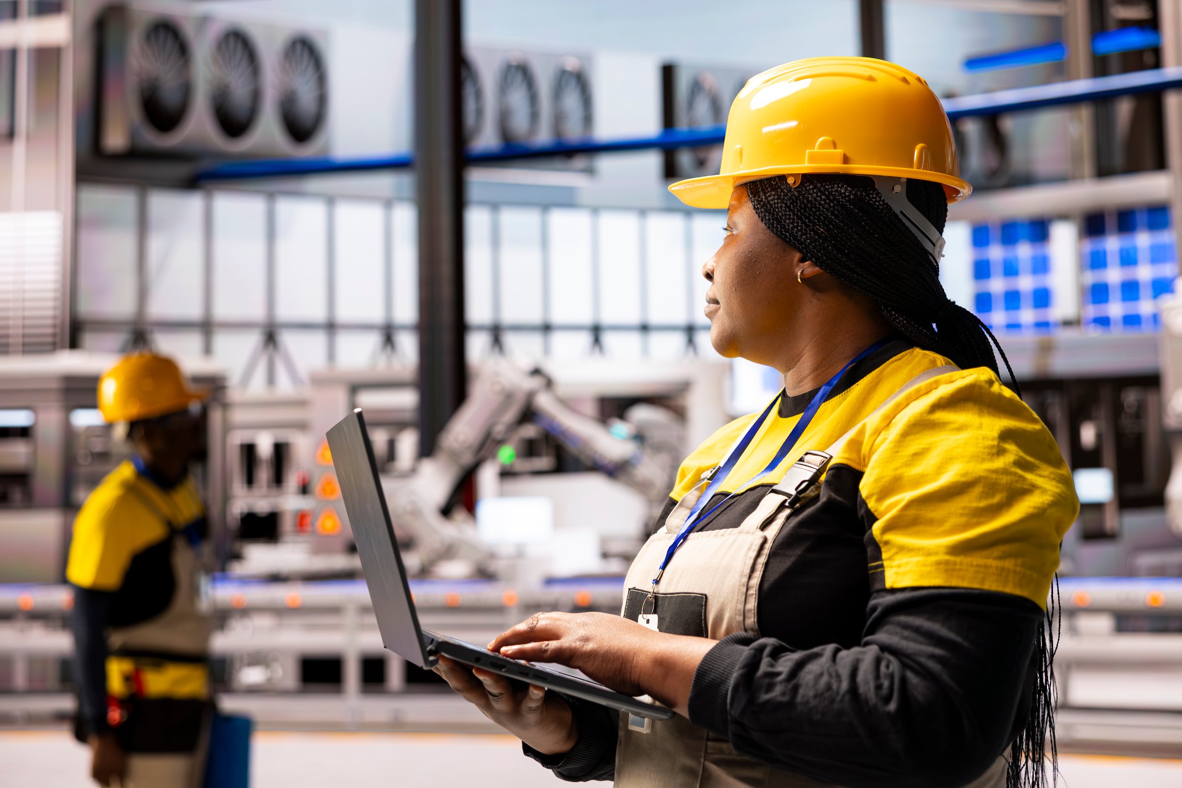 African american engineer in industrial warehouse uses laptop to monitor automated machinery. Industry 4.0 female expert controlling technological advancements in smart manufacturing facility.
