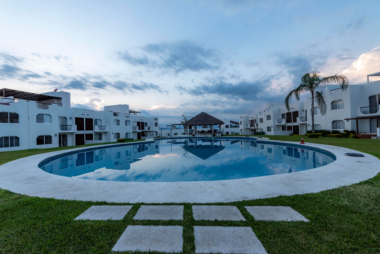 Circular pool reflecting sky, surrounded by white buildings and grass, with a thatched-roof gazebo in the background.