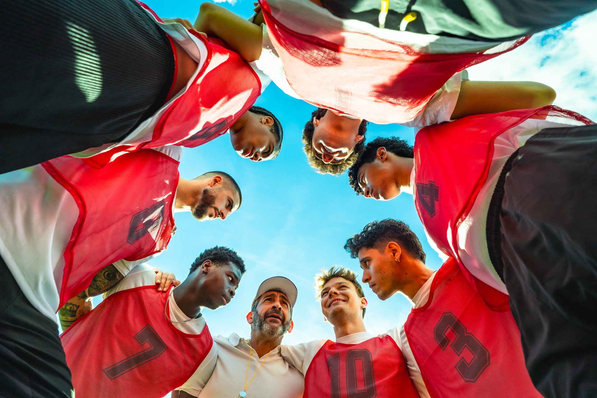 Diverse male football players and coach in a close huddle under a blue sky, arms linked, sharing strategy, motivation and team unity before a match or training session