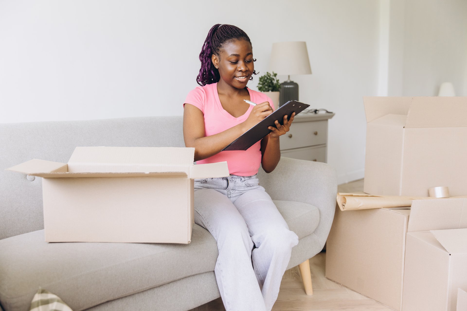 African american woman sitting on a couch, organizing and writing on a clipboard amidst moving boxes in a bright, modern living room